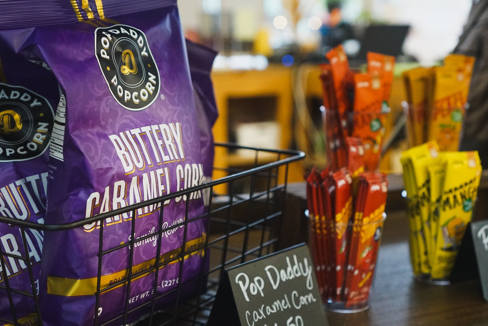 A purple bag of popcorn labeled 'Butterly Caramel Corn' in a wire basket at a snack stand, with red and yellow candy packages in the background.