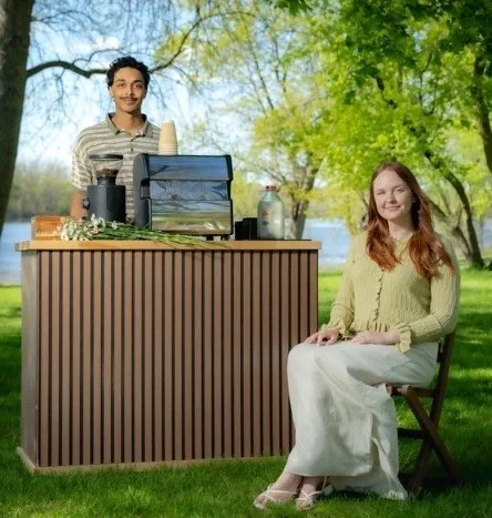 A woman sitting outdoors at a green park with a body of water in the background, smiling, with a man standing behind a food or drink stand.