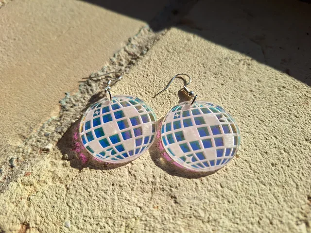 Round earrings with a cutout of a globe design, placed on a textured surface with sunlight and shadow.