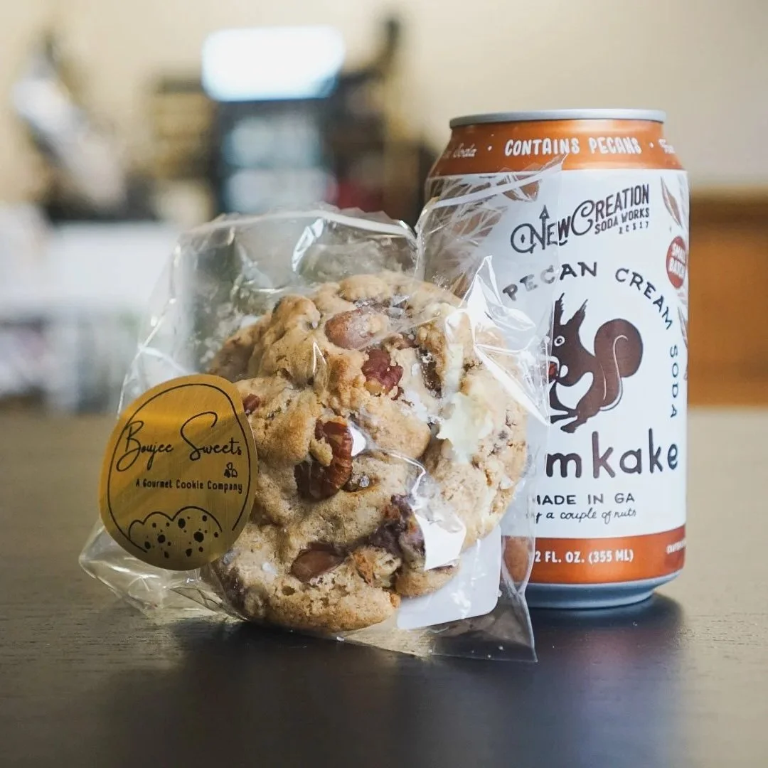 A clear plastic bag of pecan cookies with a yellow sticker reading 'Boyee Sweets, A Gourmet Cookie Company,' placed next to a can of pecan cream soda with a squirrel logo and the words 'Pecan Cream Soda' in black on a white background, on a dark surface with a blurred background.