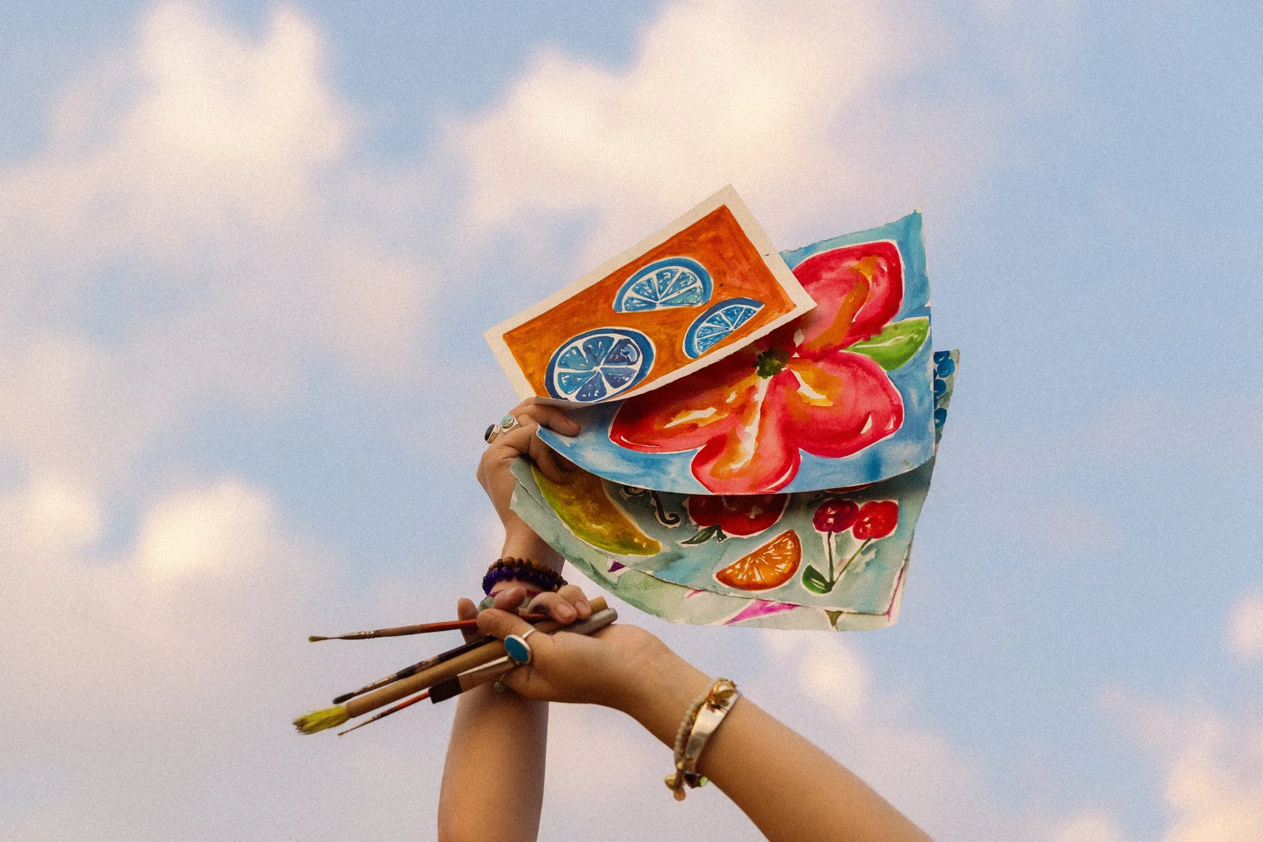 Person holding colorful paper lanterns with painted fruit and floral designs against a cloudy sky.