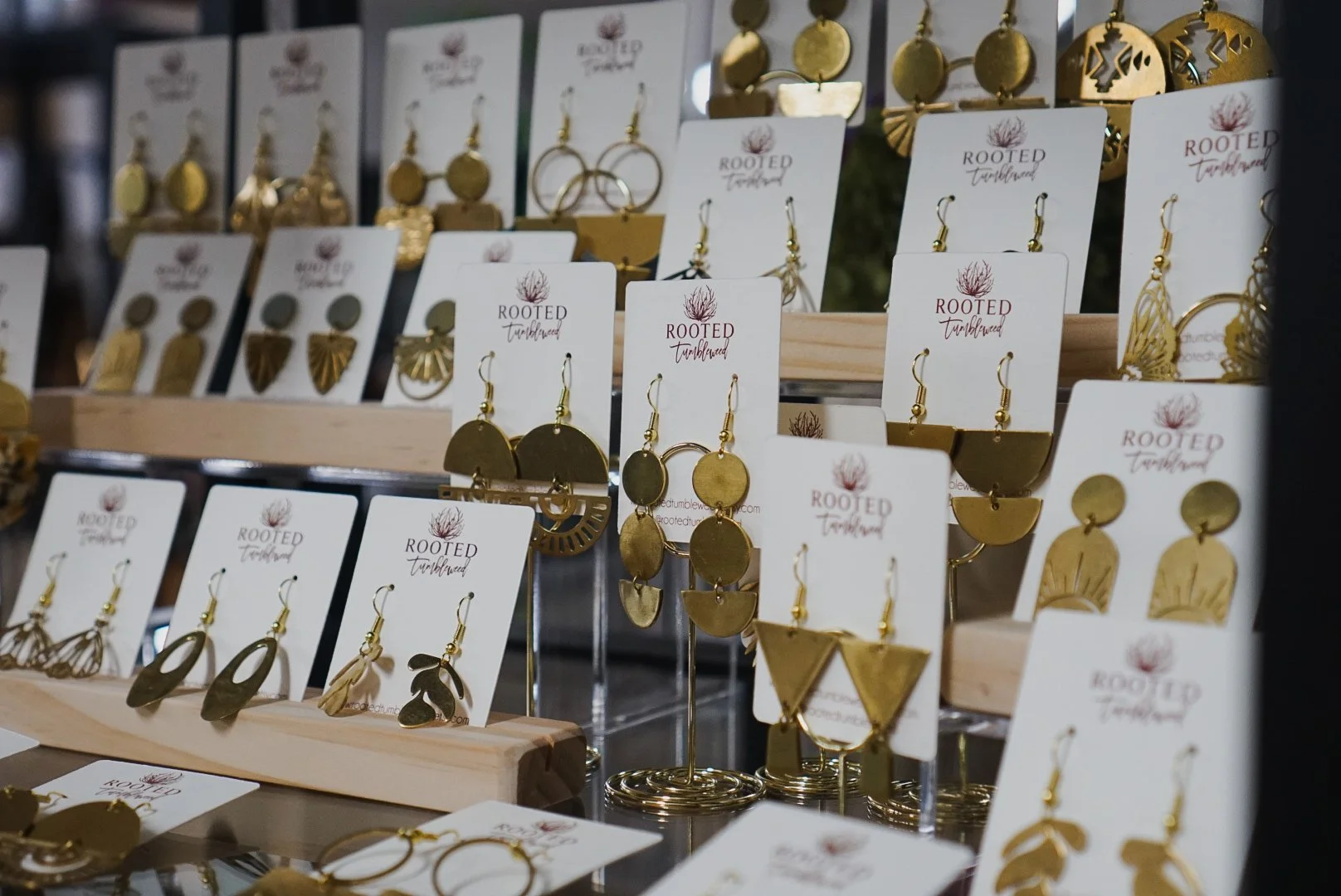 Display of gold earrings on jewelry cards with 'Rooted Turbulence' branding at a craft or jewelry market.