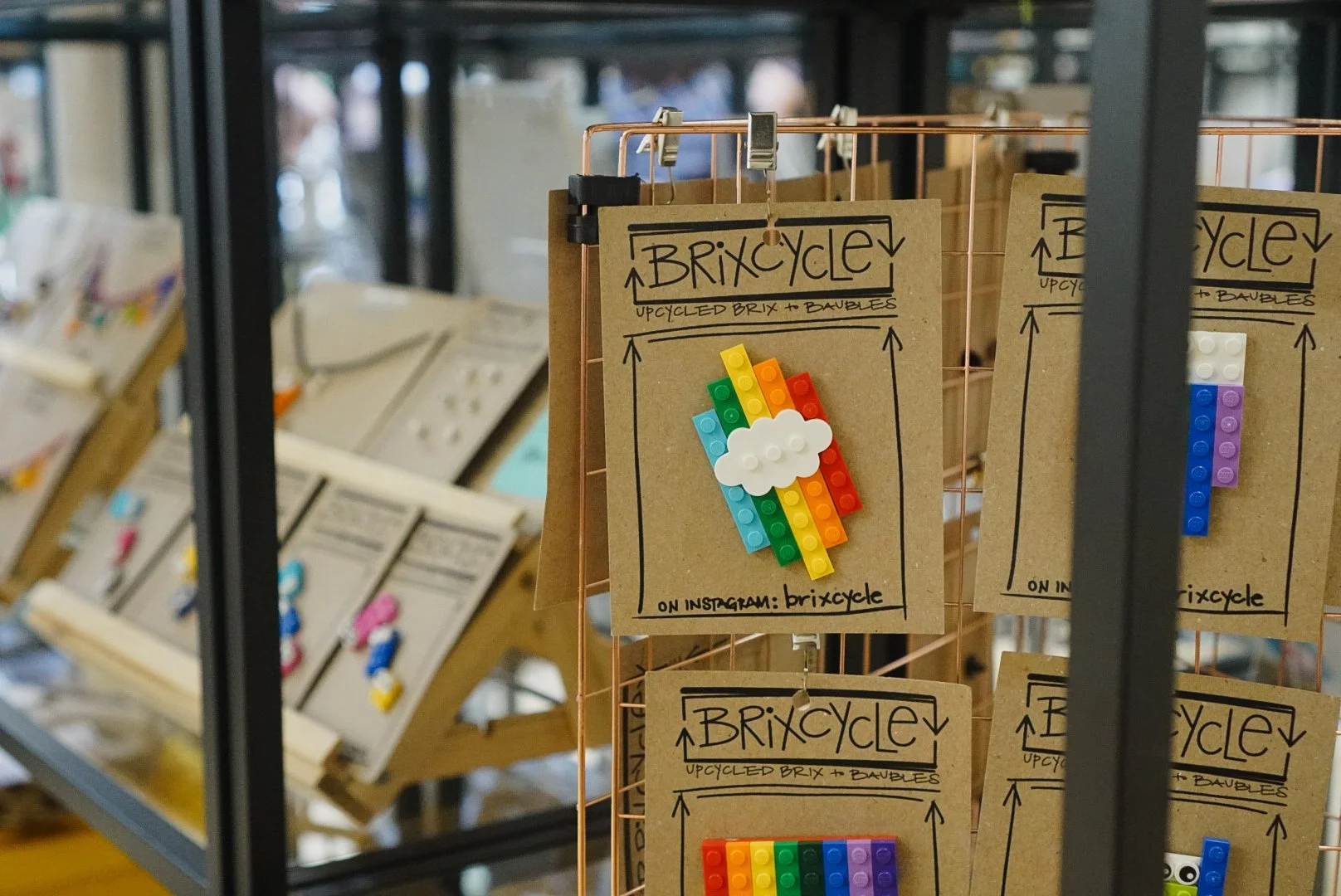 Display of upcycled Lego art on cardboard cards, with the word 'BRIXCYCLE' and an Instagram handle, featuring a rainbow and cloud Lego piece.