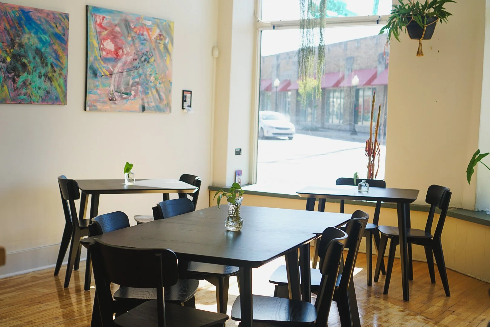 Interior of a cafe or restaurant with black tables and chairs, decorated with small potted plants, large abstract paintings on the wall, and a large window letting in natural light.