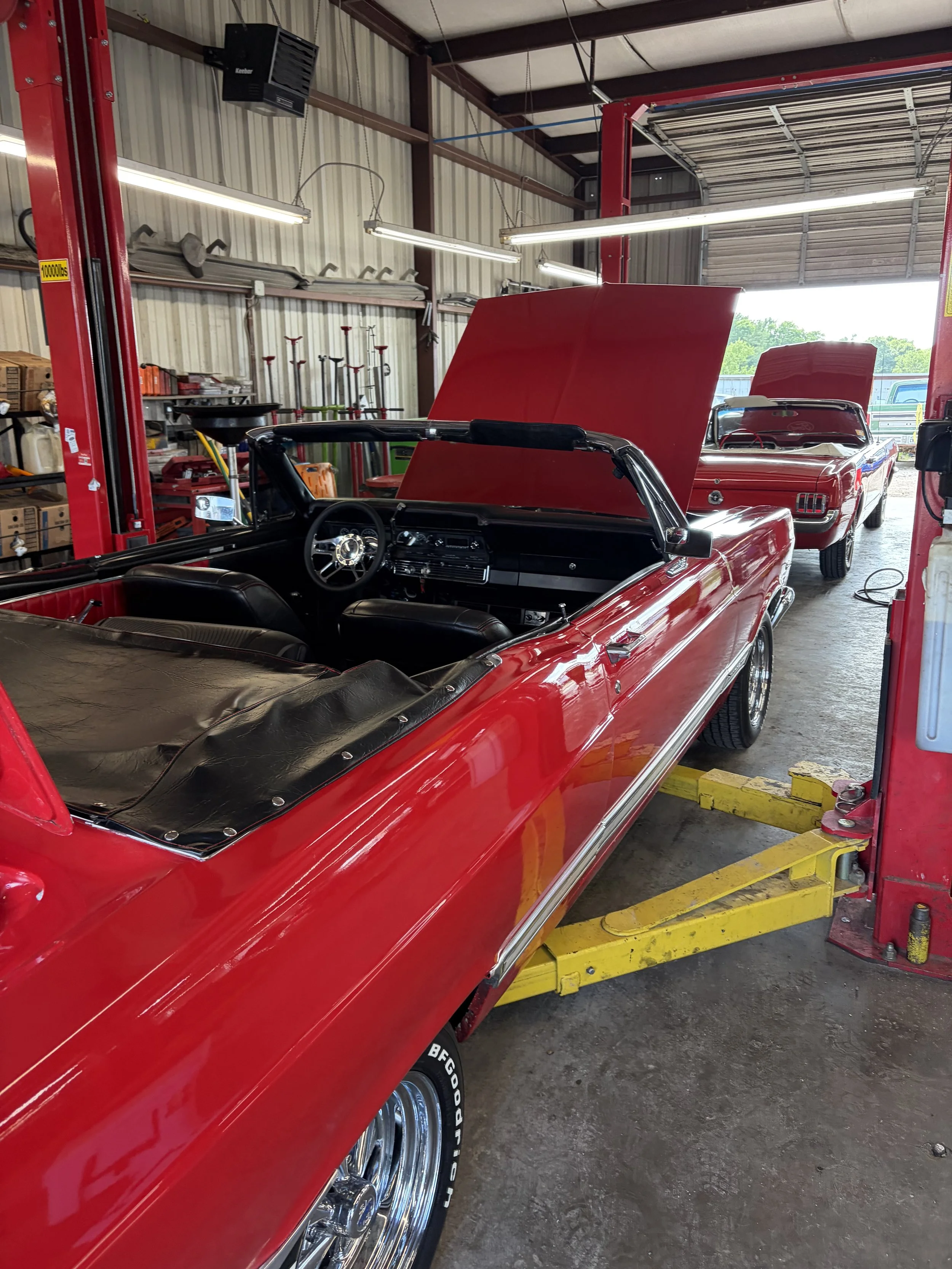 Inside a garage with two red classic cars, one with its hood open. The garage has metal walls, fluorescent lighting, and various tools and equipment on shelves.