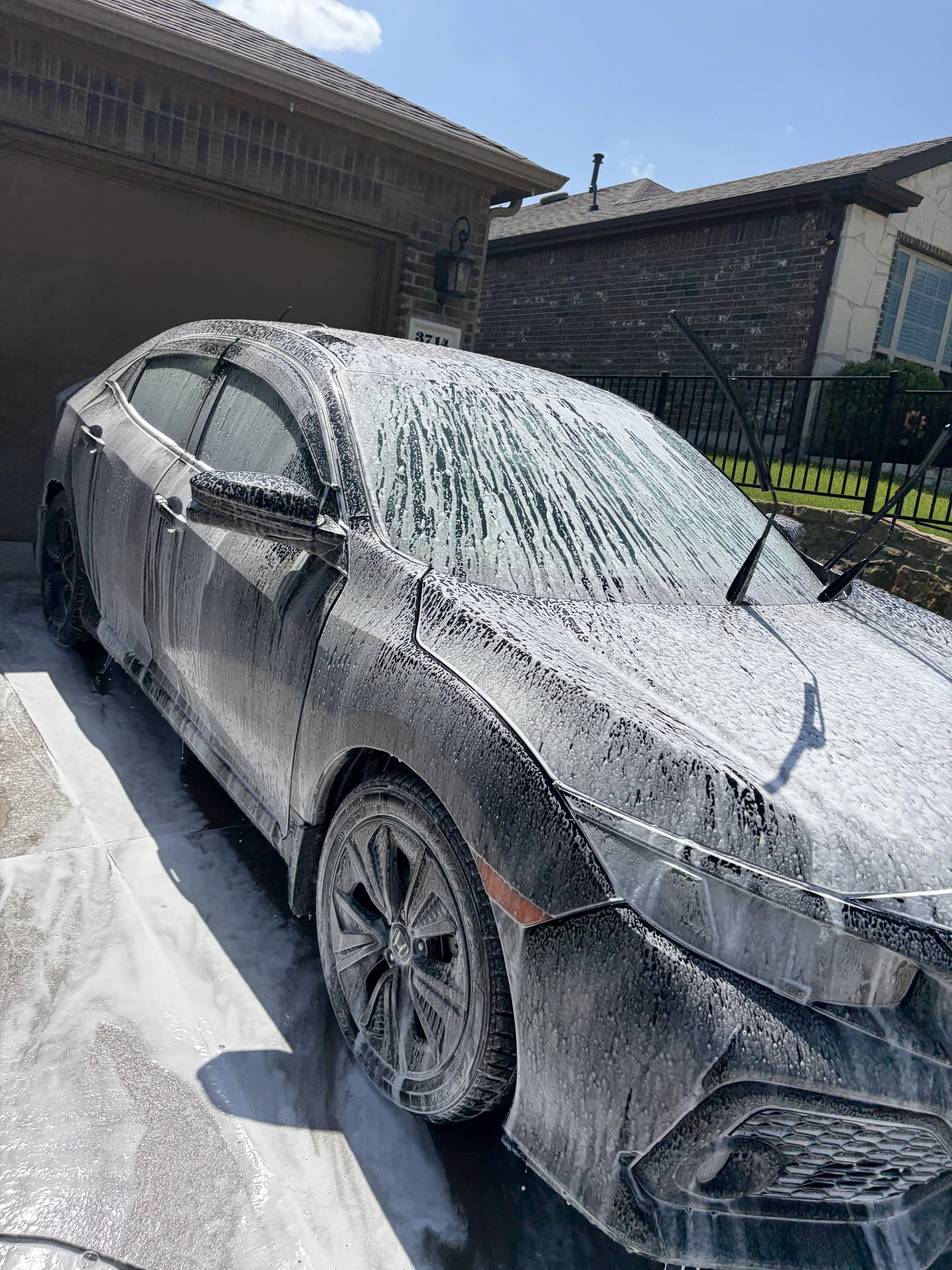 A black car covered in white soap and foam during a car wash, with soap bubbles on the windshield, roof, and sides, parked on a driveway outside a house with brick and stone walls.