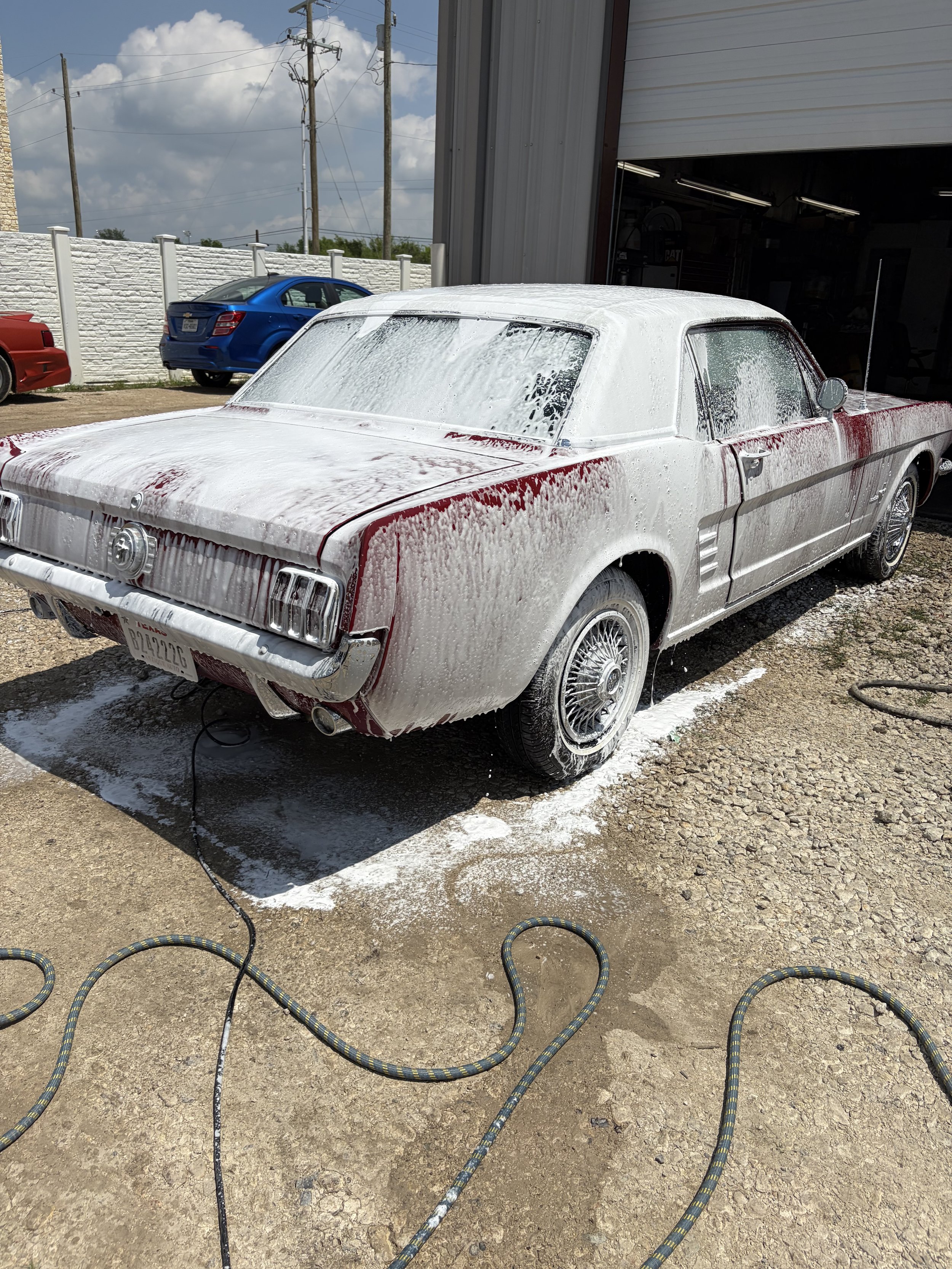 A vintage red and white car being washed with soap, surrounded by hoses on a gravel ground outside a garage, with a white brick wall, a blue car, and utility poles in the background.
