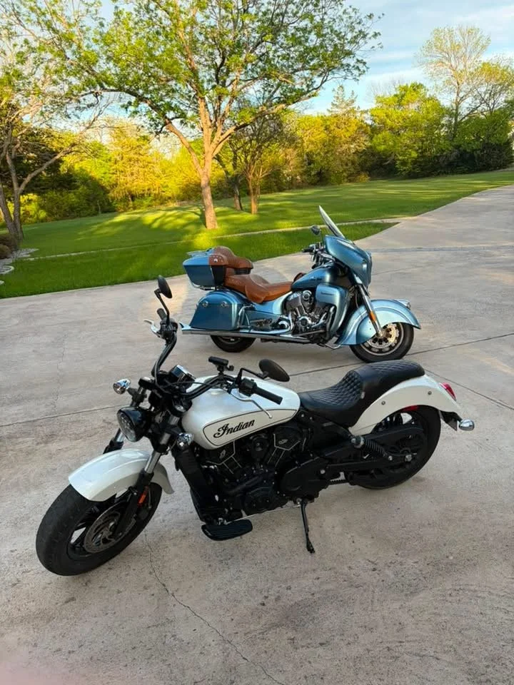 A white Indian motorcycle and a blue touring motorcycle parked on a concrete driveway with green grass and trees in the background.