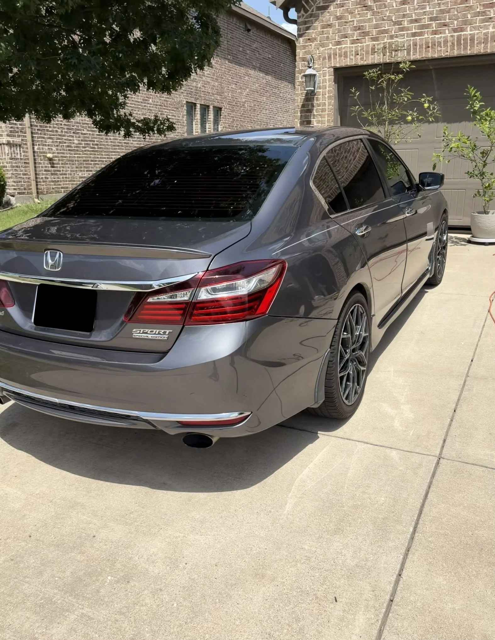 Gray Honda sedan parked in a driveway with brick house wall in the background, surrounded by a tree with green leaves and a potted plant.