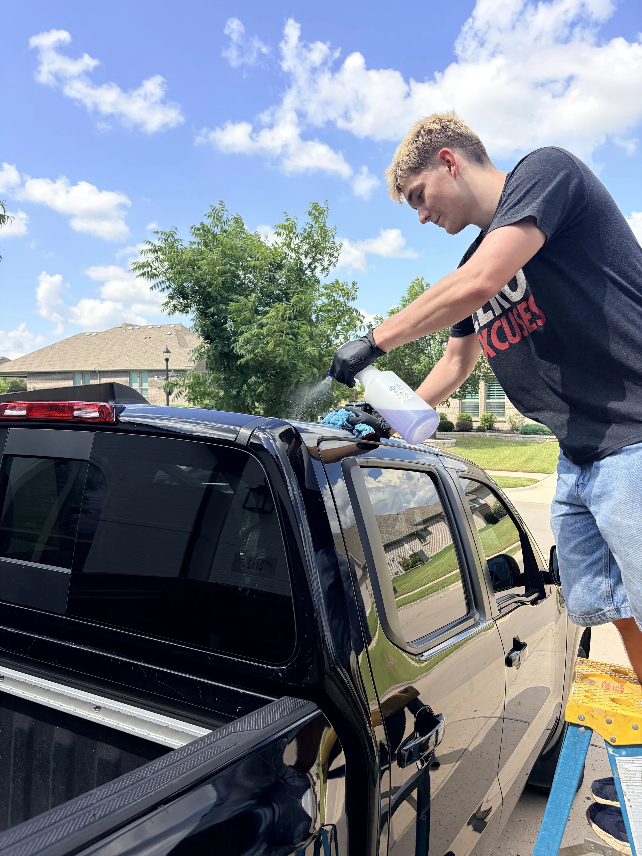 A young man with blond hair wearing a black shirt and jeans is detailing a black truck outdoors on a sunny day, using a spray bottle and cloth.