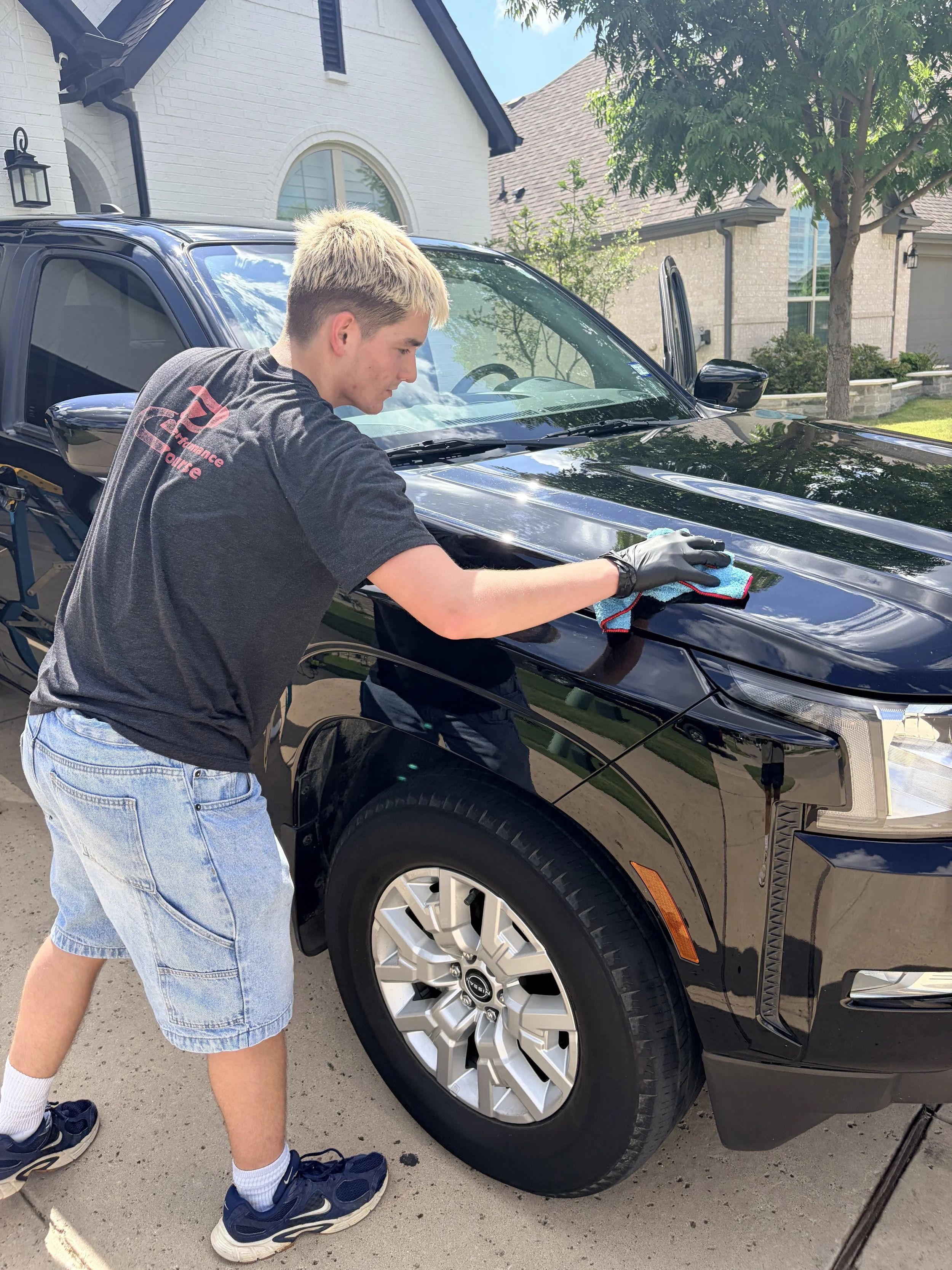 A young boy with blond hair, wearing a black T-shirt with red and pink text, denim shorts, white socks, and blue sneakers, is cleaning the hood of a shiny black pickup truck with a cloth in a residential driveway.