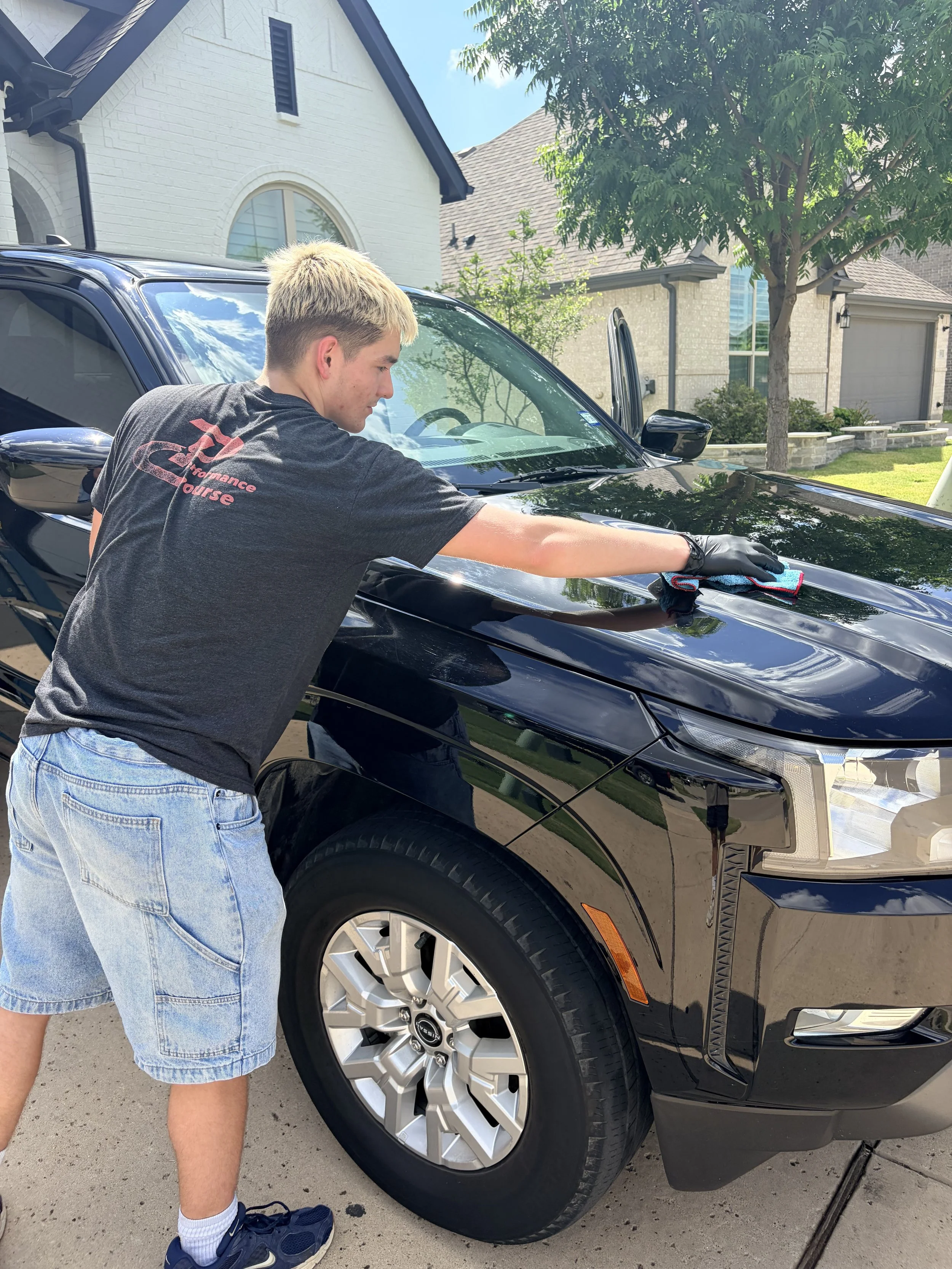 A young person with blond hair, wearing a black shirt, denim shorts, and blue sneakers, is cleaning the hood of a black car with a microfiber cloth in a driveway of a suburban house.