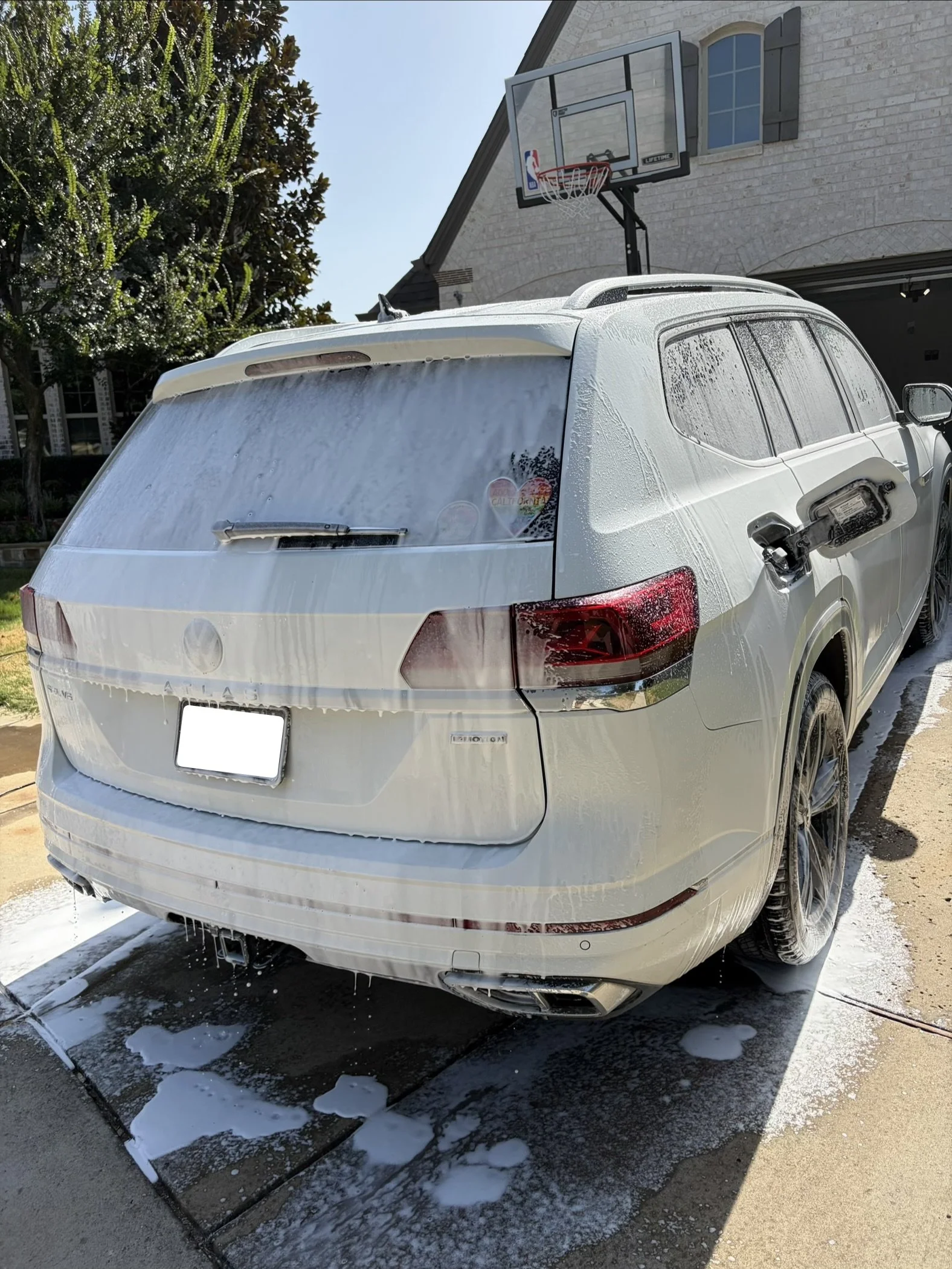 A white SUV is being washed with soap and water, with foam on the rear window and body, parked outside a house with a basketball hoop in the driveway.