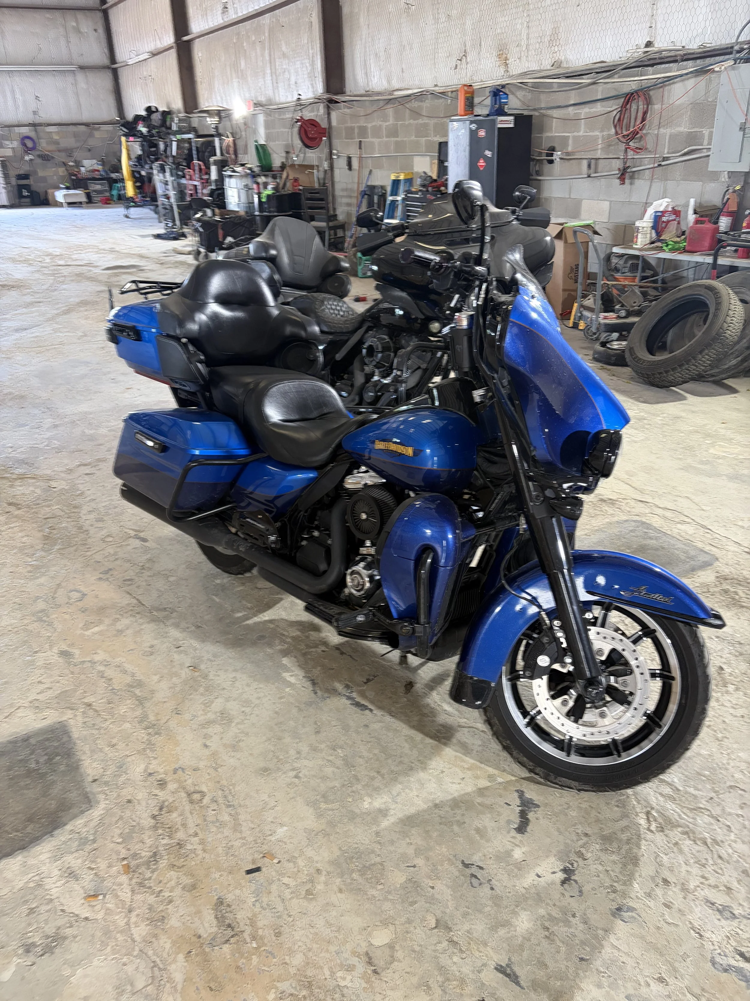 Blue touring motorcycle with black leather seats and large front windscreen parked inside a workshop with concrete floors and cinderblock walls, surrounded by tools, tires, and workshop equipment.