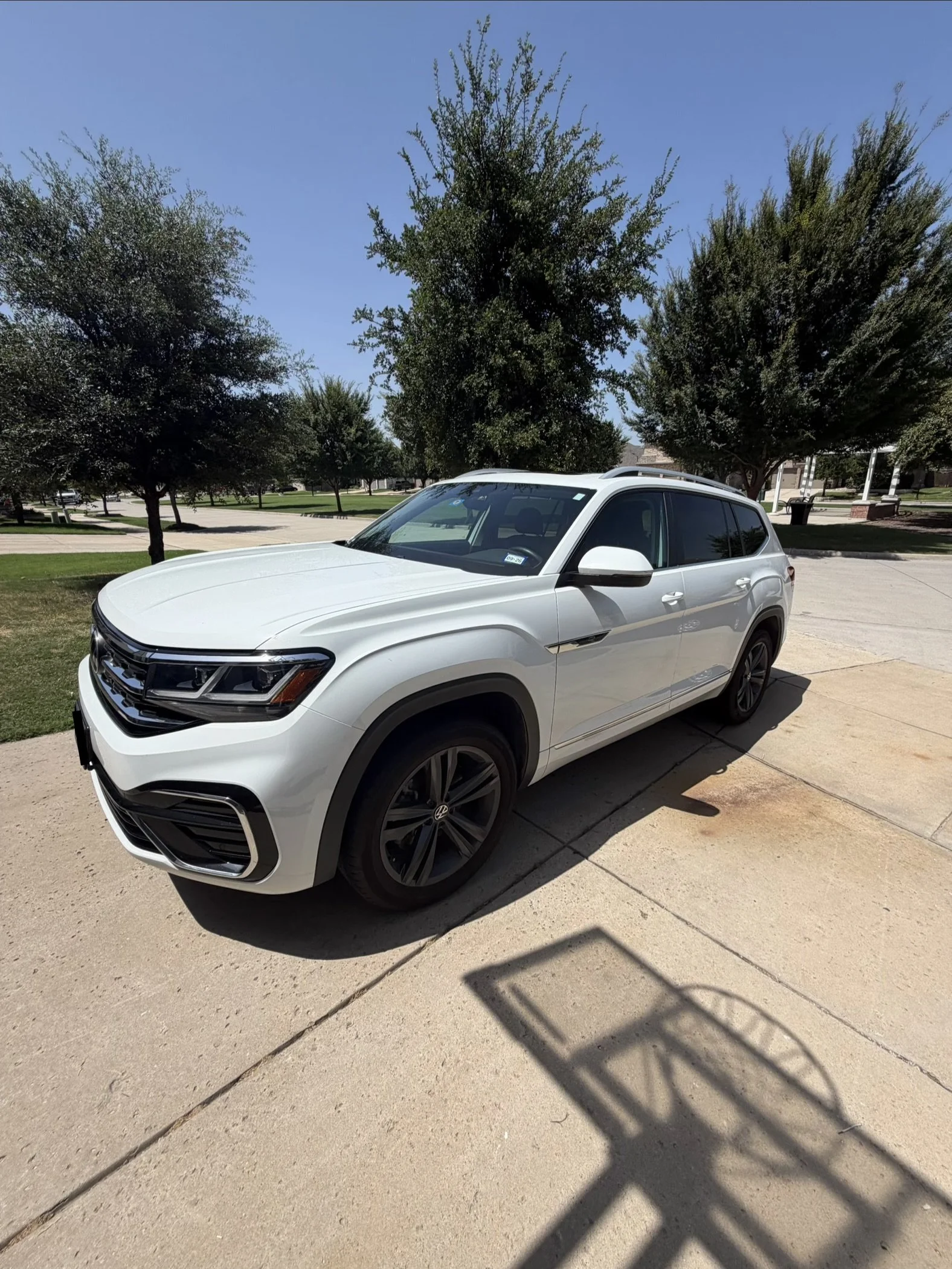 A white SUV parked on a concrete driveway under trees in a residential neighborhood with houses and a park in the background.