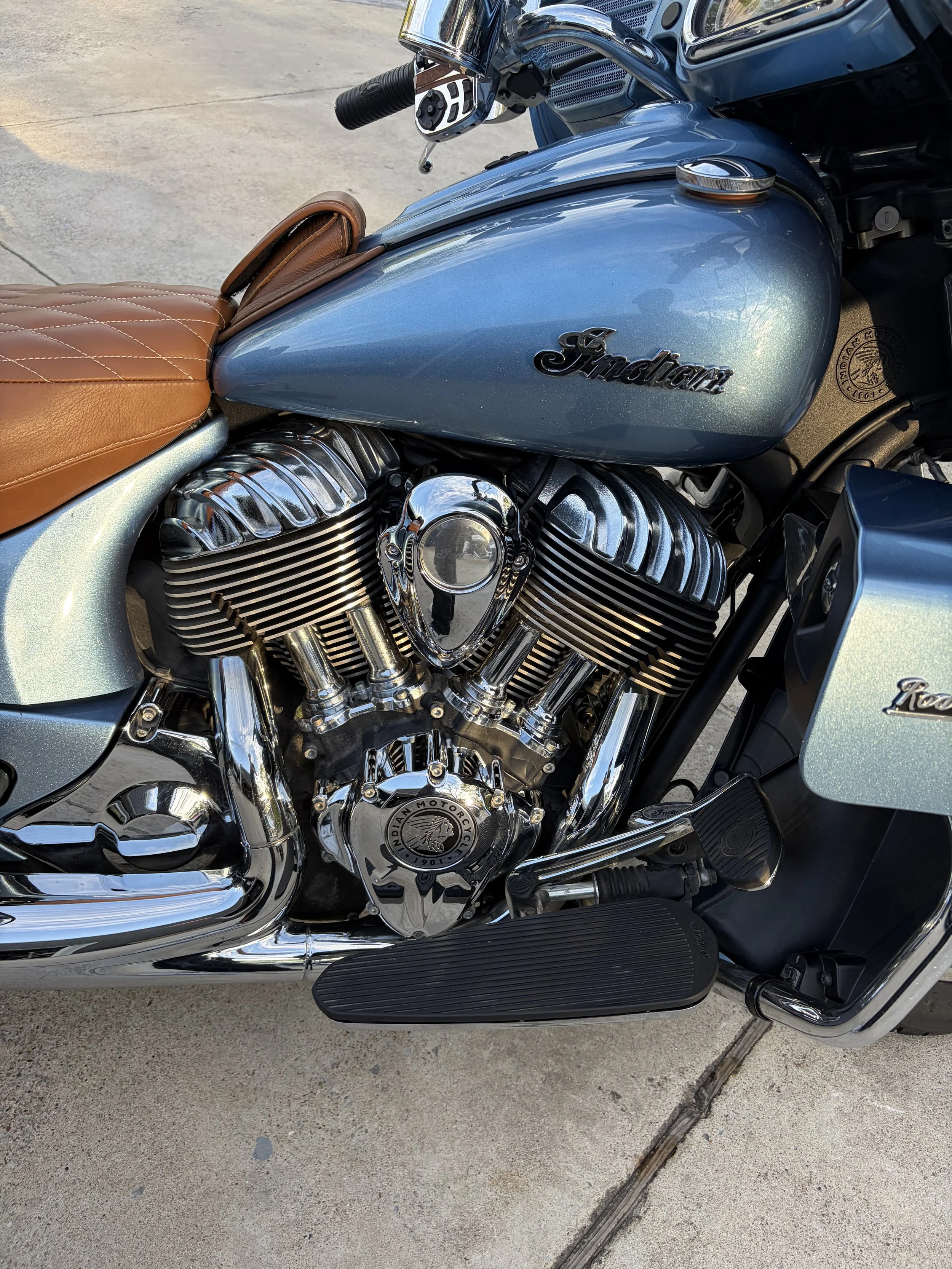 Close-up view of a Harley-Davidson motorcycle's chrome engine, blue fuel tank with 'Indian' logo, brown leather seat, and black footrest, parked on a concrete driveway.