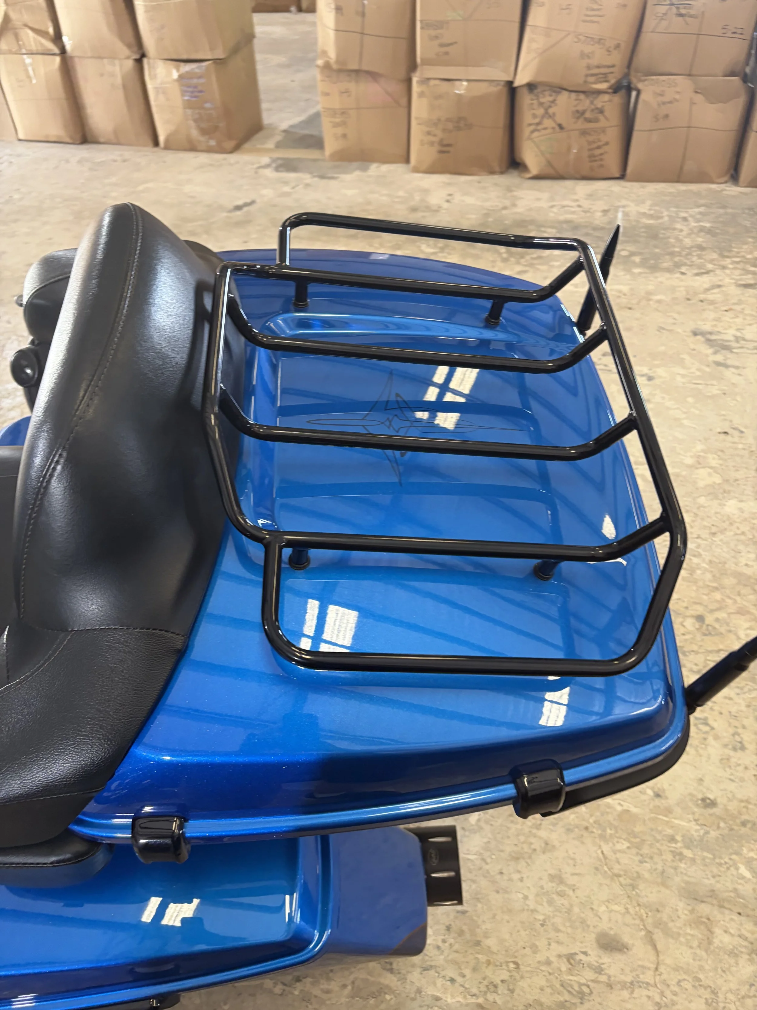 A blue mobility scooter with a black seat and a black metal basket attached to the back, parked on a concrete floor in front of large cardboard boxes.