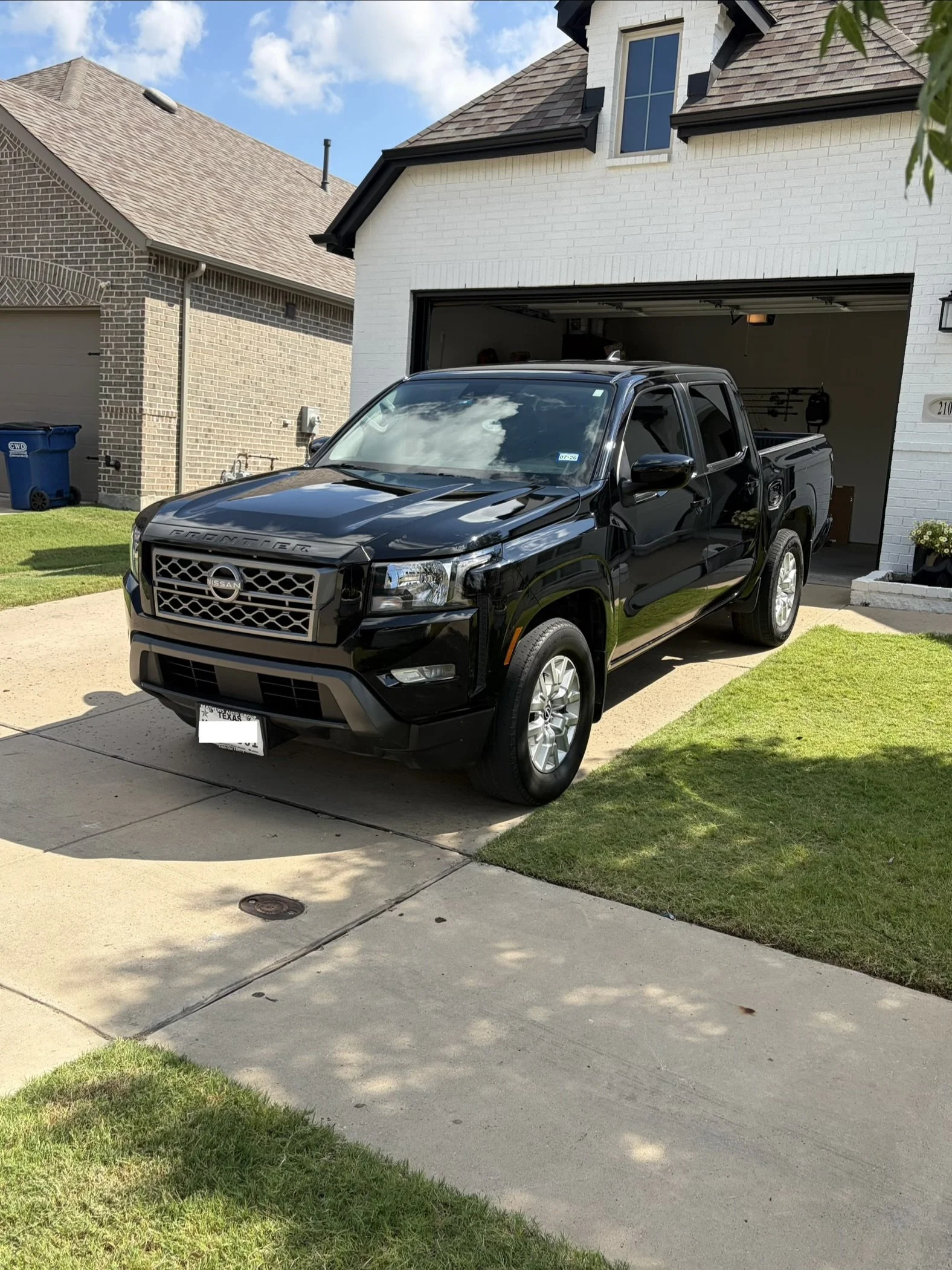 A black Nissan Frontier pickup truck parked in front of a white brick house with an open garage door. The house has a brown shingle roof, a small dormer window, and black trim. A grassy lawn and sidewalk are visible in the front yard, with a blue recycling bin on the left side.