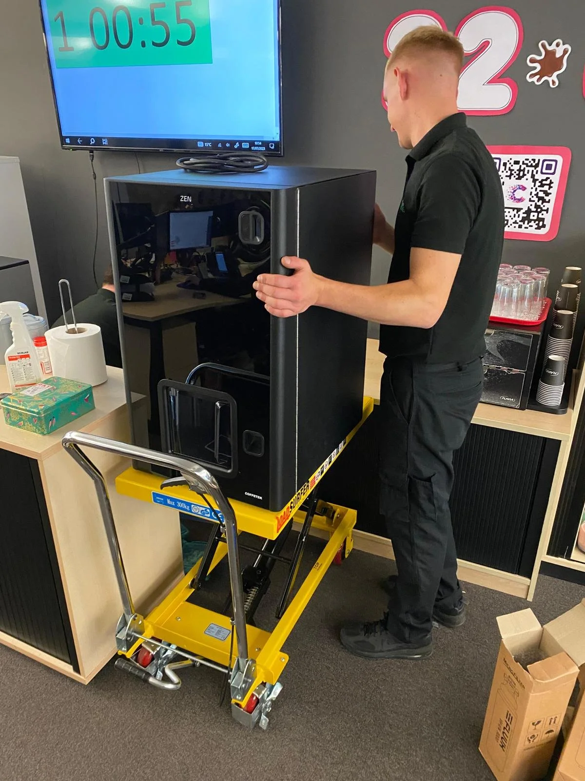 A person is moving a large black appliance, using a yellow platform lift in an office setting. There is a wall-mounted digital timer display, and various office supplies and refreshments are visible on nearby furniture.