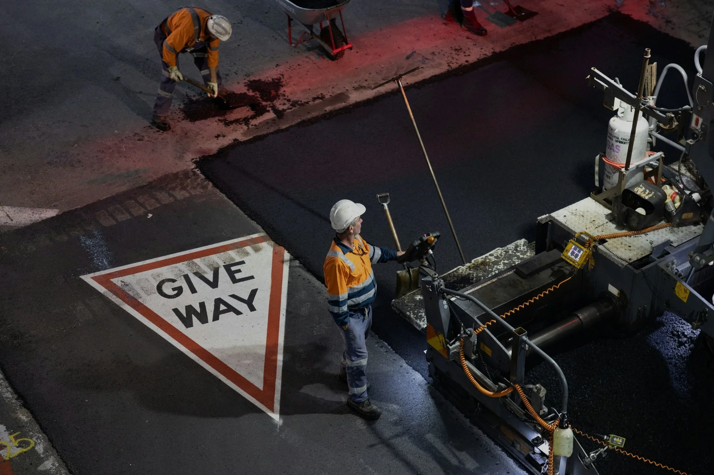 Construction workers paving a road at night, with a 'Give Way' sign on the street and industrial equipment around.