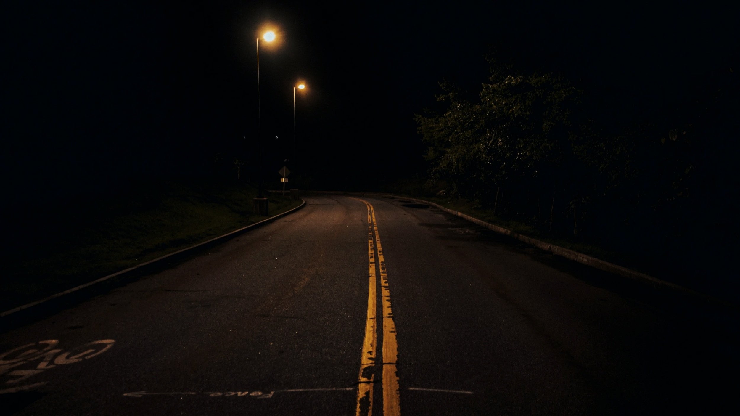 Dark street at night with streetlights casting a glow, two yellow lines in the middle, and trees on the side.