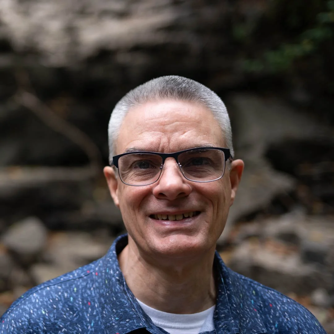 A smiling middle-aged man with short gray hair, glasses, and a blue patterned shirt, standing outdoors with a blurred natural background.