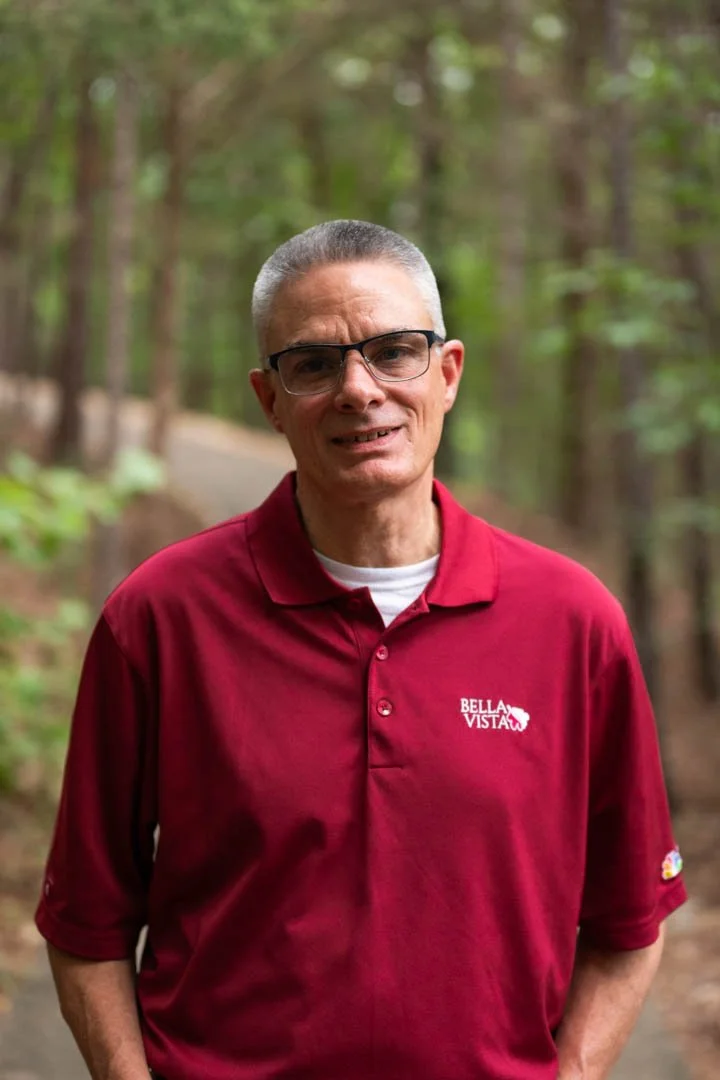 A man with glasses and short gray hair standing on a forest trail, wearing a red polo shirt with a 'Bella Vista' logo.
