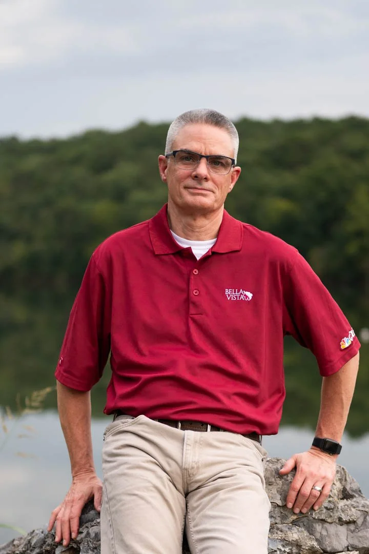 A middle-aged man wearing glasses, a maroon polo shirt with 'BELLA VISTA' logo, and beige pants, sitting on a rock outdoors near a body of water with a green wooded background.