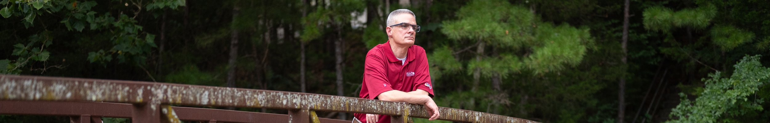 A man with short gray hair and glasses leaning on a rusty railing outdoors, with a background of green trees.