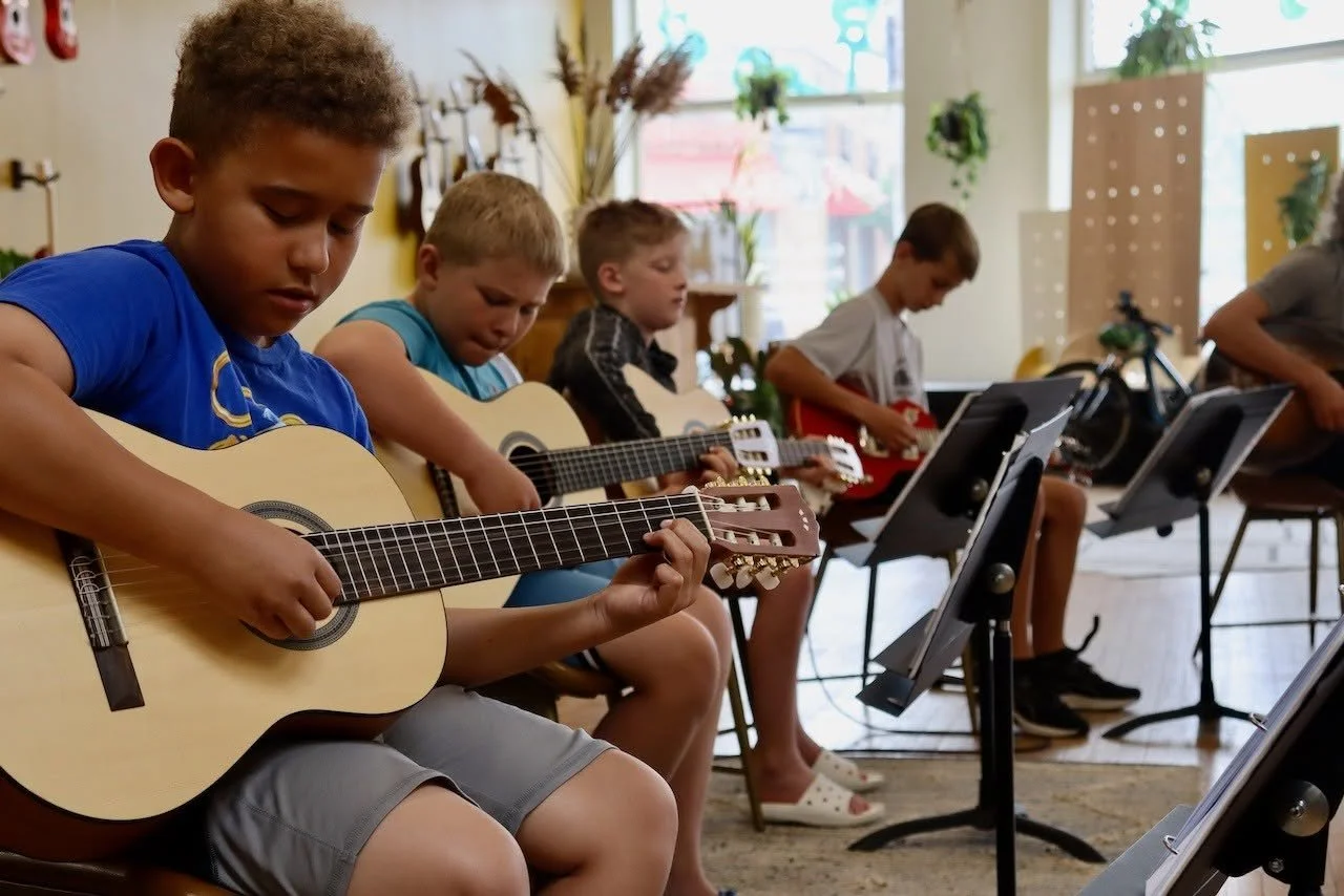Children sitting in a music classroom playing acoustic guitars, with sheet music stands in front of them and a decorated, well-lit room with windows in the background.