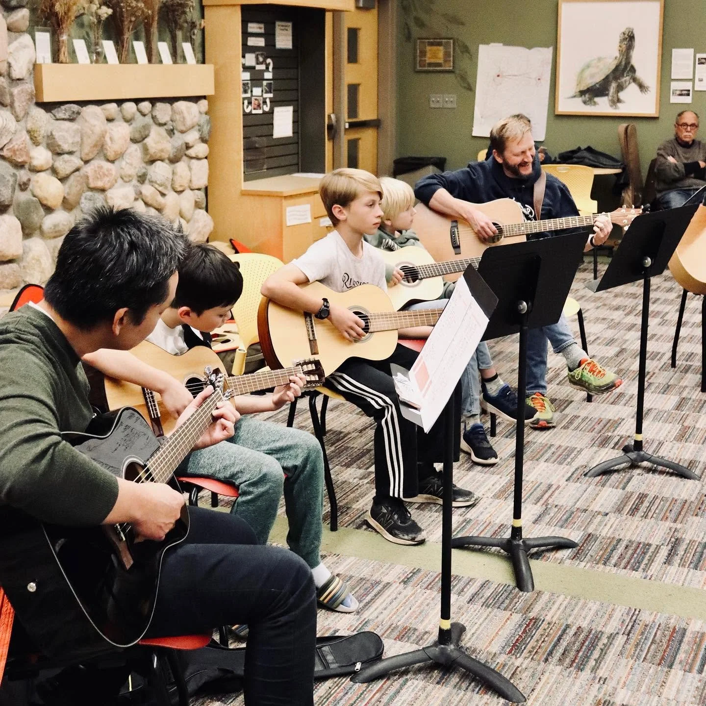 A group of children and adults playing acoustic guitars during a music session in a room with stone walls and artwork on the walls.