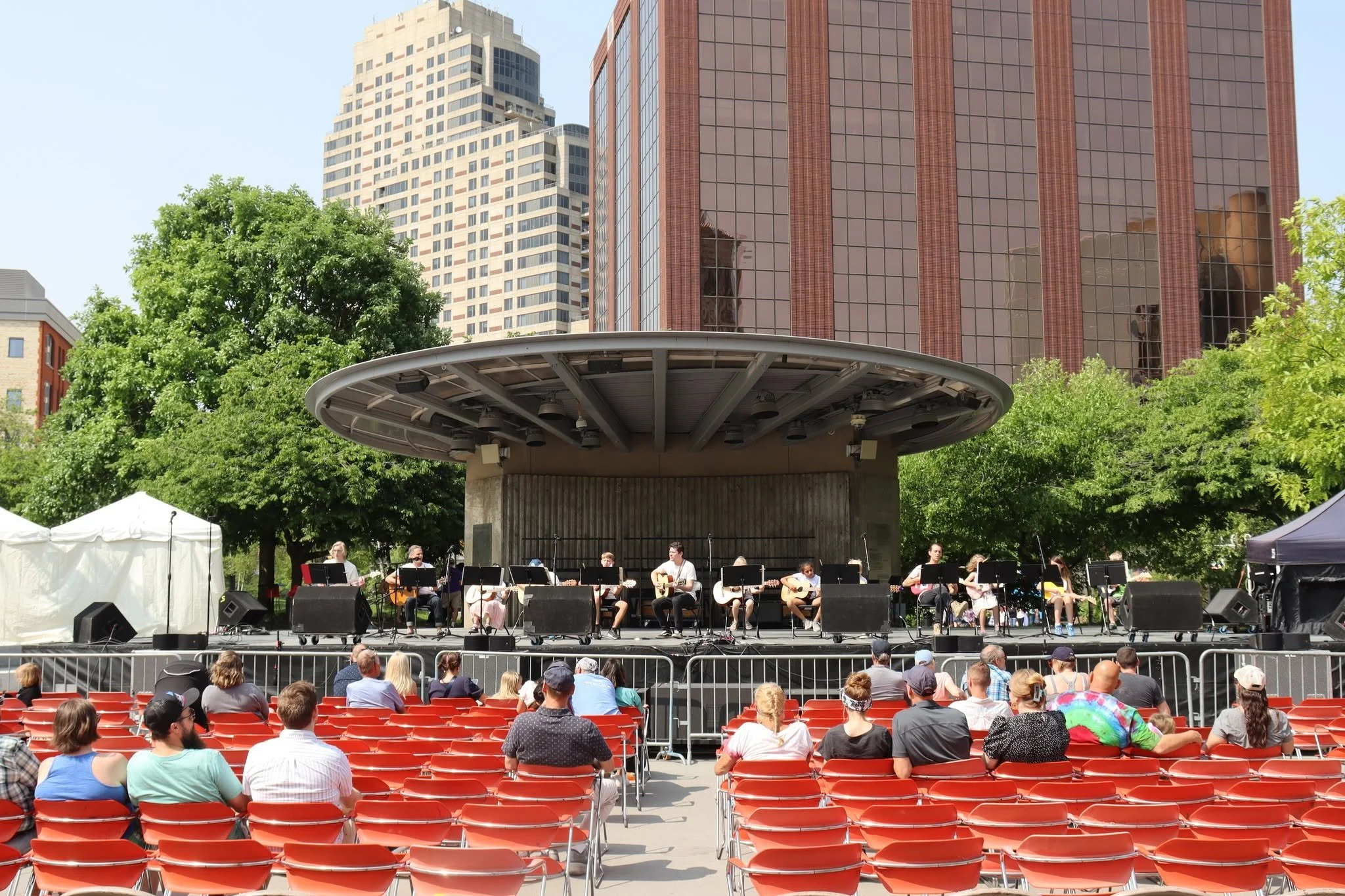 Outdoor concert stage with musicians playing guitars, audience seated in red chairs, city buildings in background, trees surrounding the stage.