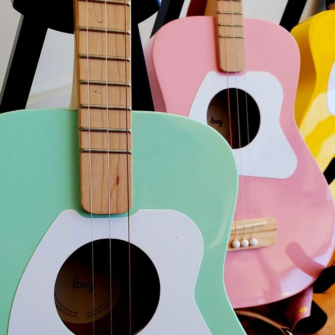 Three colorful toy guitars with wooden necks and strings, in green, pink, and yellow, lined up against a black chair.