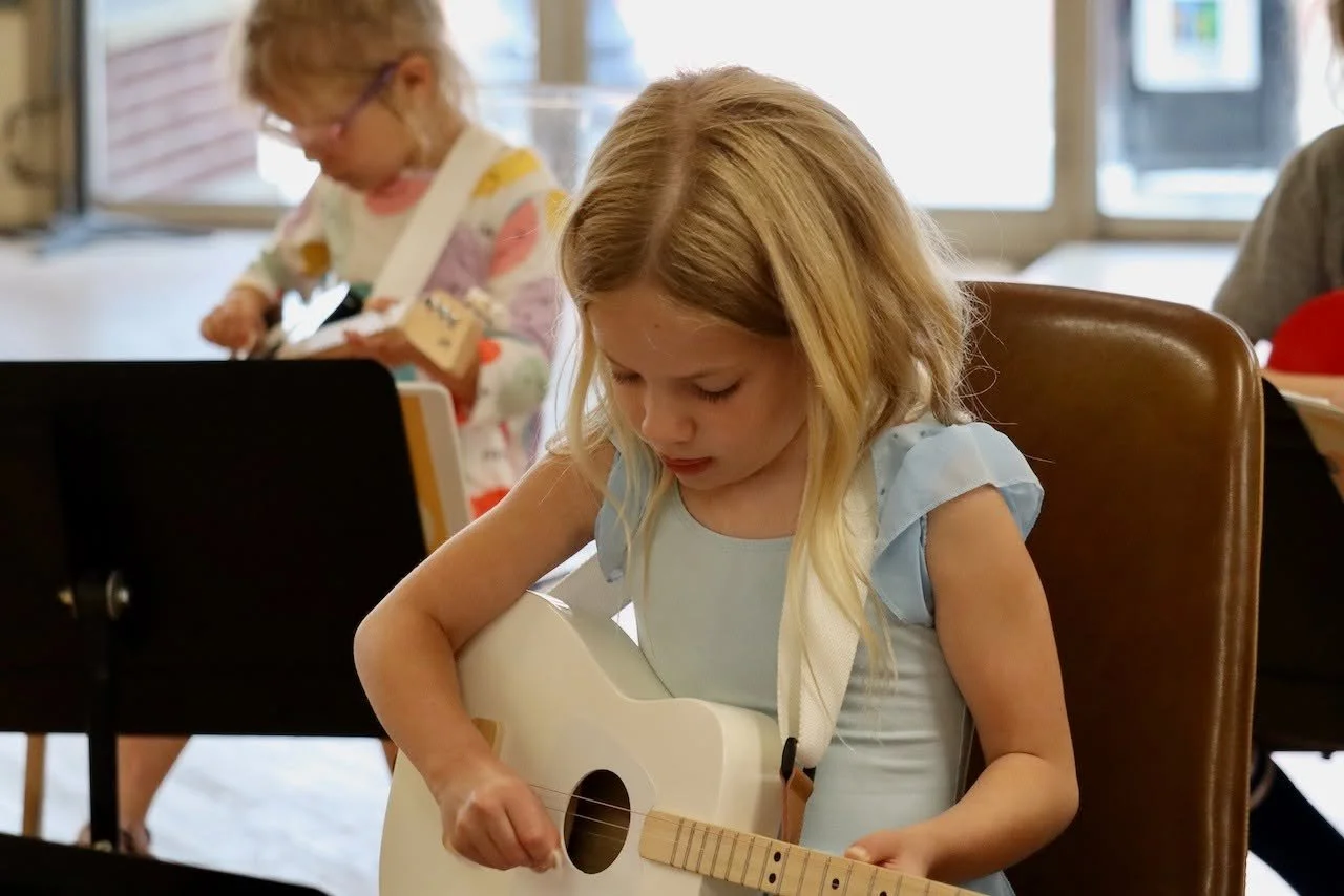 A young girl with blonde hair playing a white guitar during a music class with other children in the background.