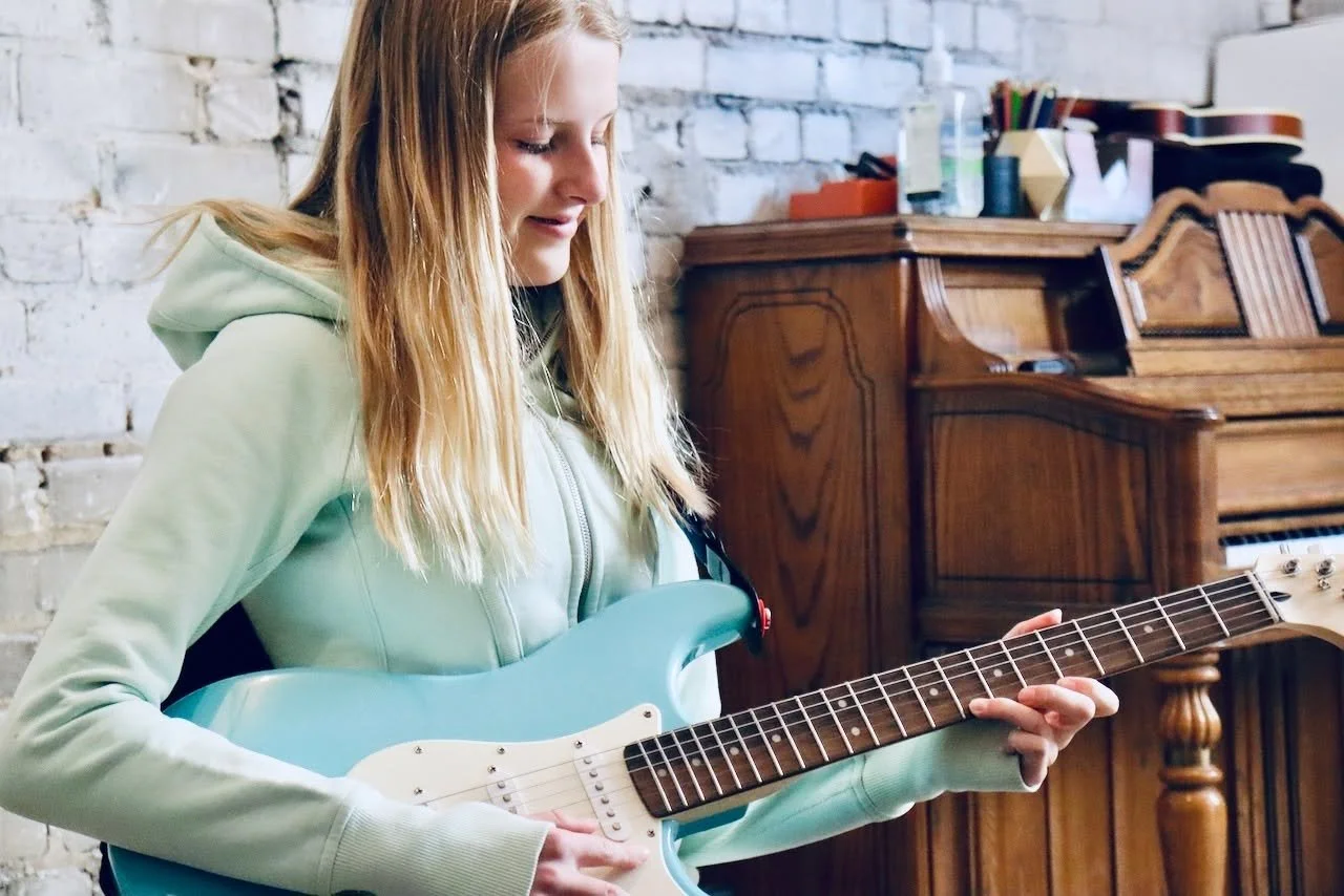 A girl with long blonde hair playing a light blue electric guitar in a room with a brick wall and wooden furniture.