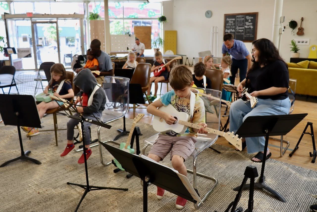 Children and adults playing guitars and ukuleles in a music class or rehearsal space, with music stands and a casual, cozy room interior.