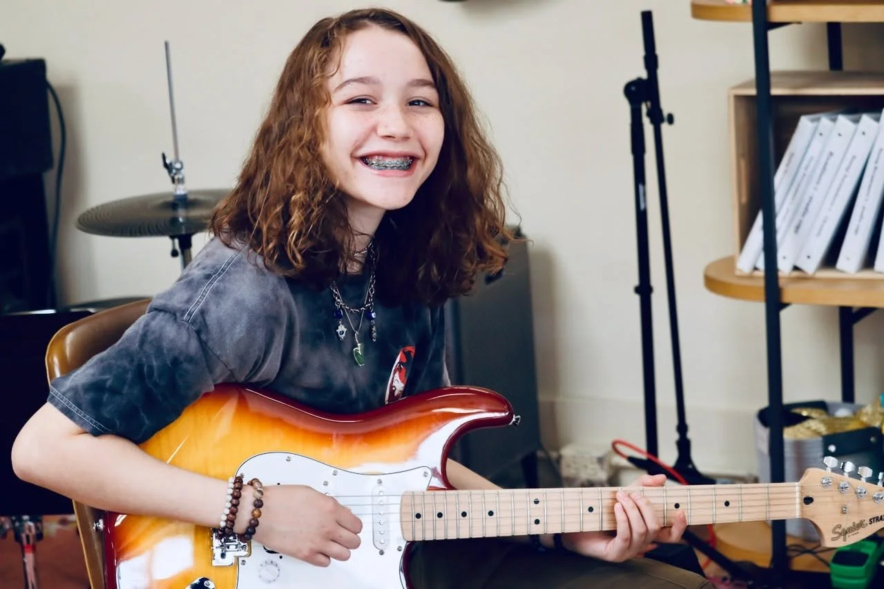 Young girl with curly hair smiling and playing an electric guitar in a room.