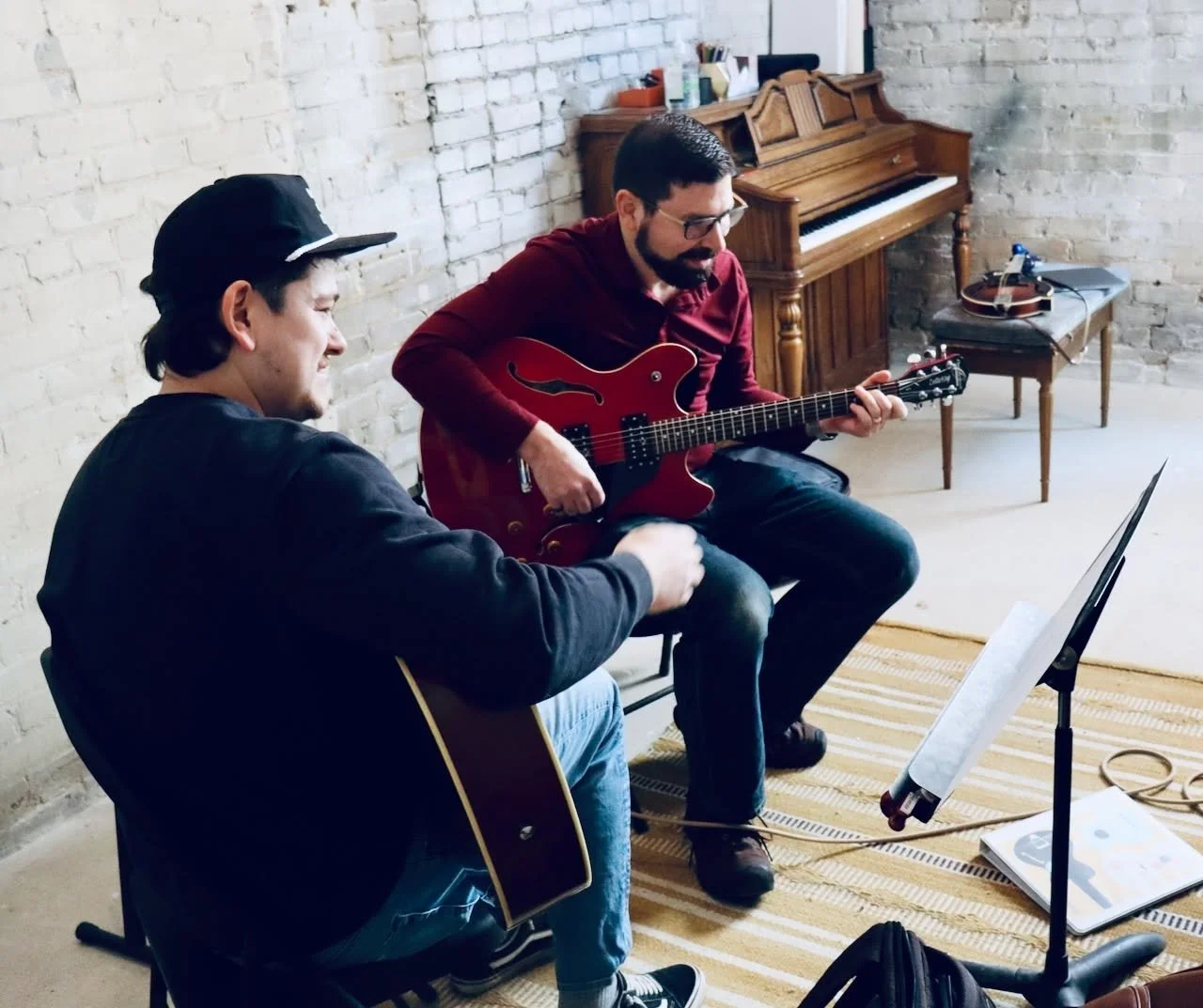 Two men playing guitars in a room with a brick wall. One is sitting and playing an acoustic guitar, while the other is sitting and playing an electric guitar. There is a music stand with sheet music or a notebook nearby, and a piano and a small table with a guitar in the background.