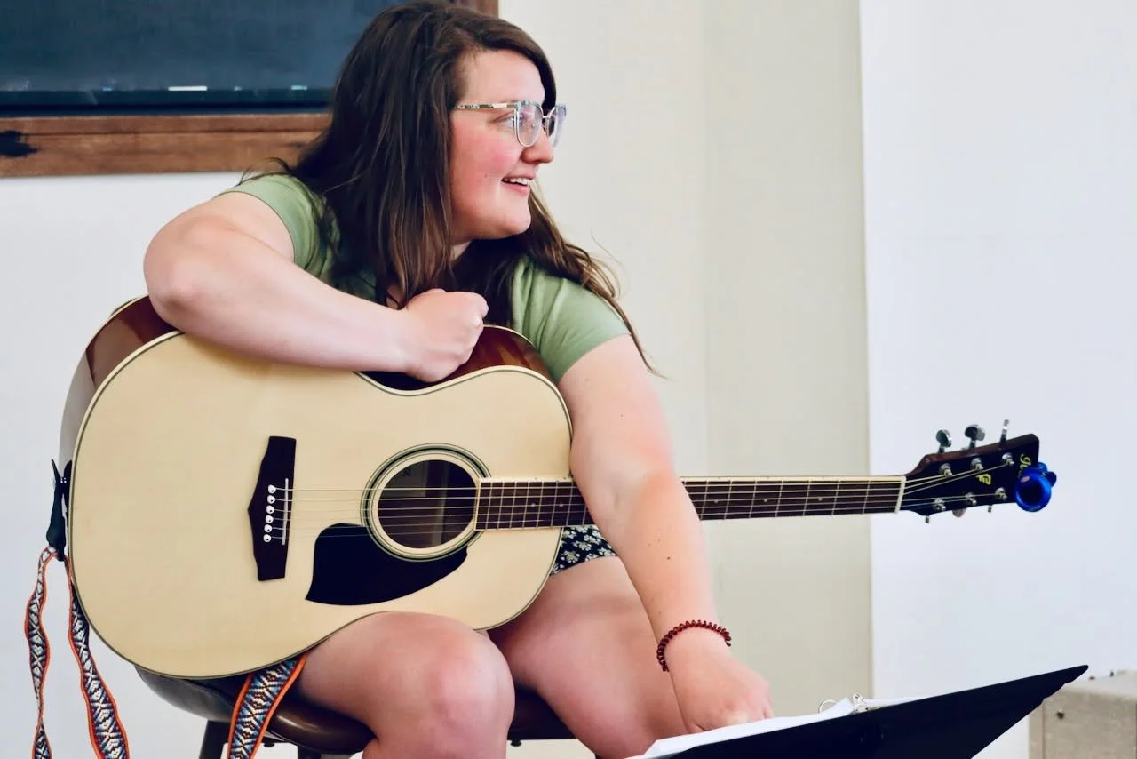 A woman with glasses, wearing a green t-shirt, sitting and playing an acoustic guitar.
