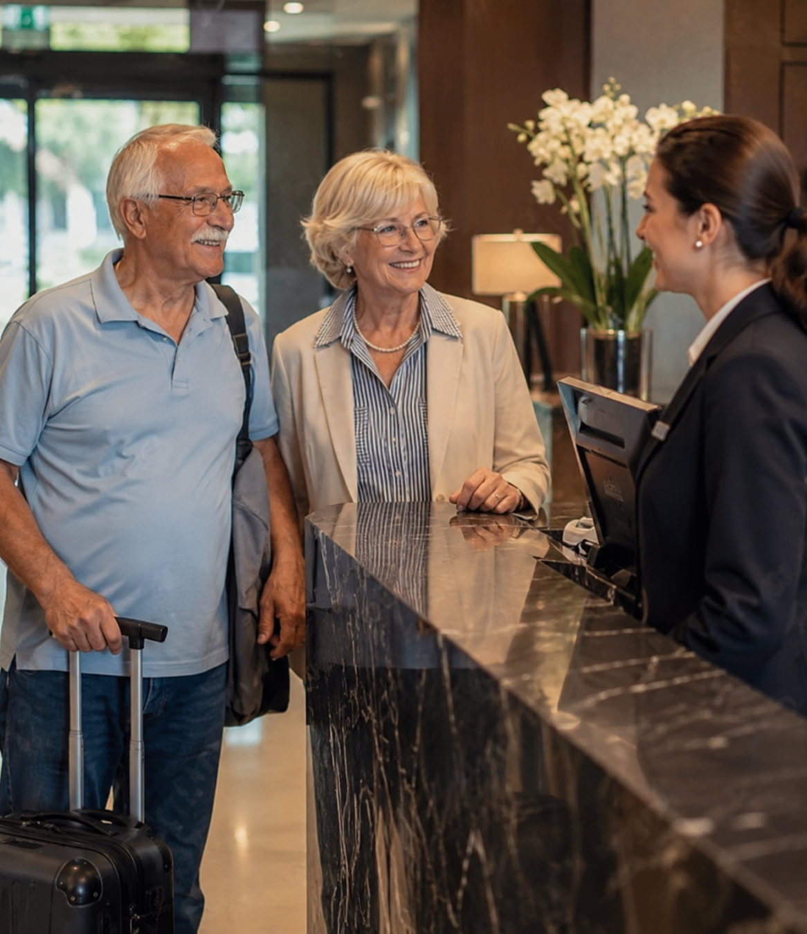An elderly man with white hair and glasses, carrying a small rolling suitcase, is talking to a woman and a hotel receptionist at the hotel lobby reception desk. The woman has blonde hair and is smiling, and the receptionist is dressed in a dark uniform and is holding a tablet.