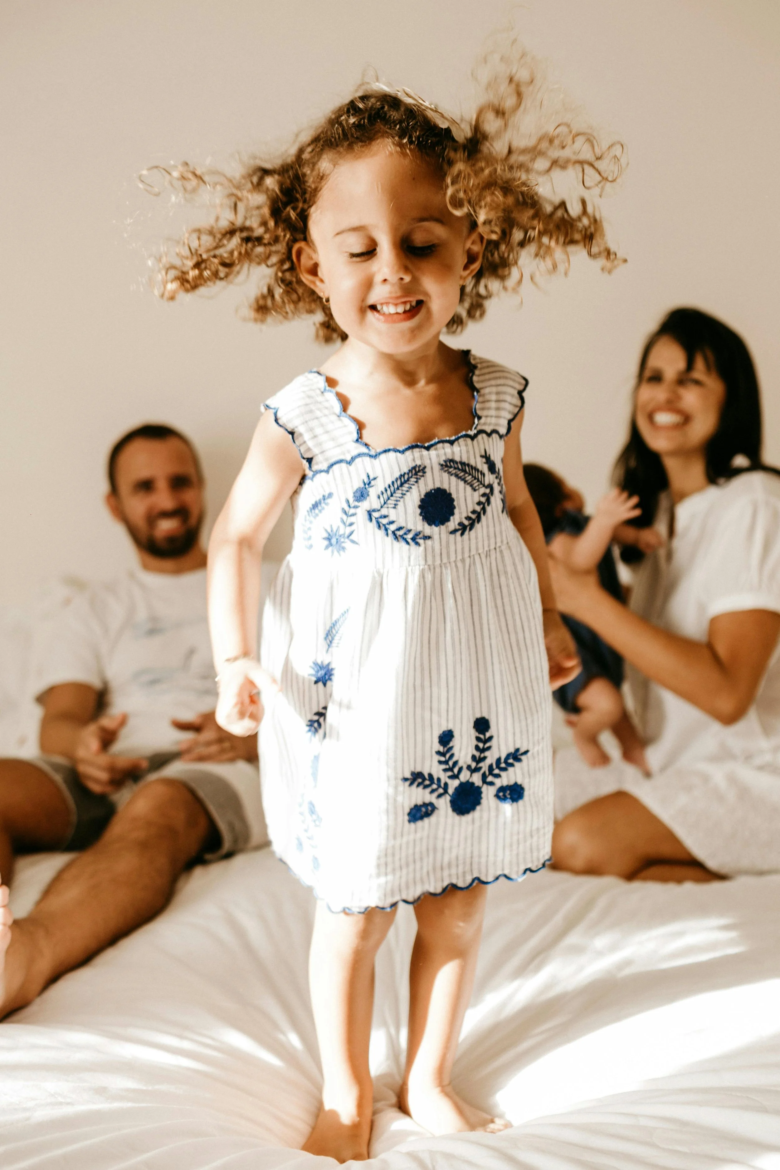 smiling happy toddler with curly hair jumping on a bed with parents smiling in background