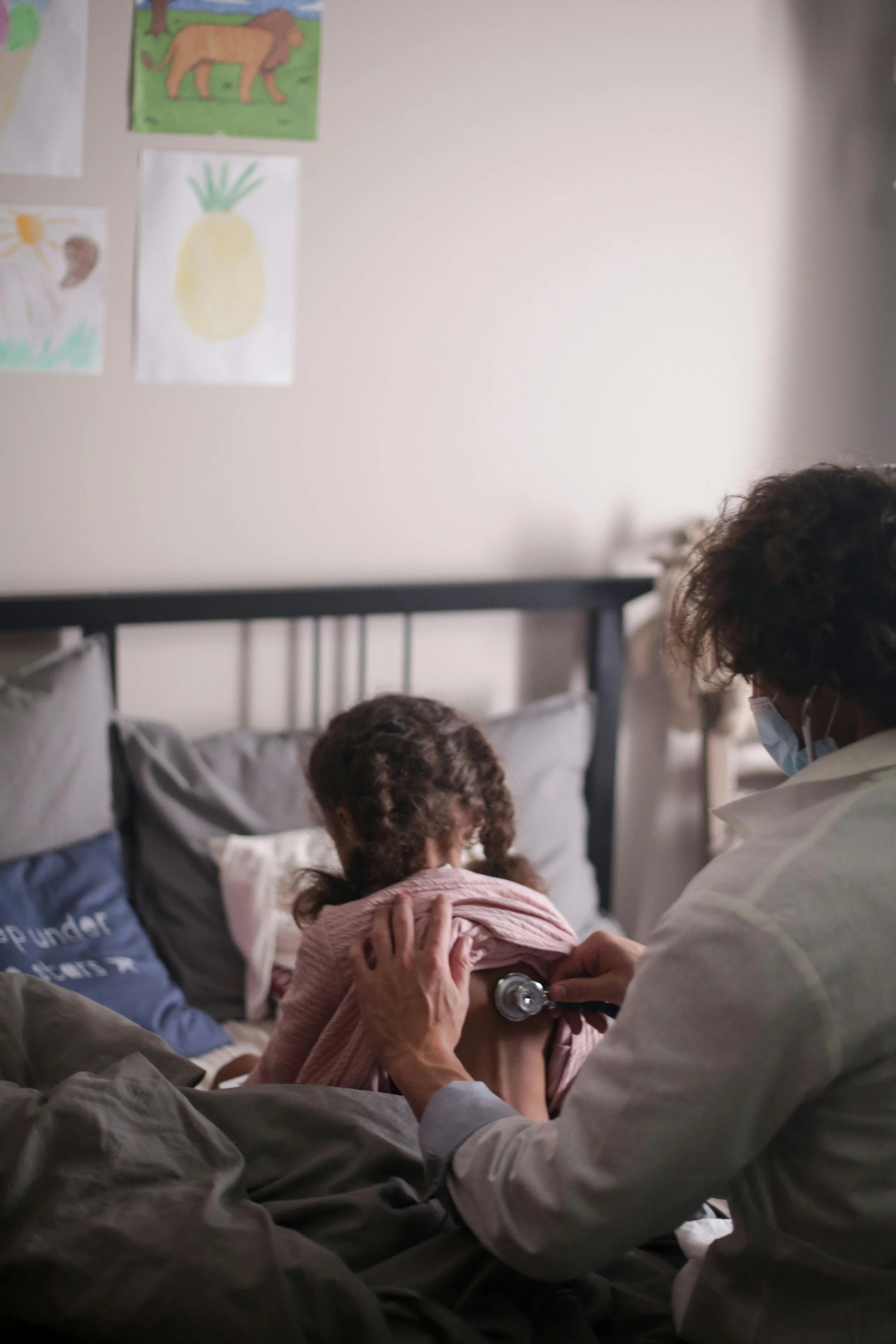 A healthcare professional in a white coat and face mask checks a young girl's heartbeat with a stethoscope. The girl is sitting on a bed with gray sheets, and there are children and colorful drawings of animals on the wall in the background.