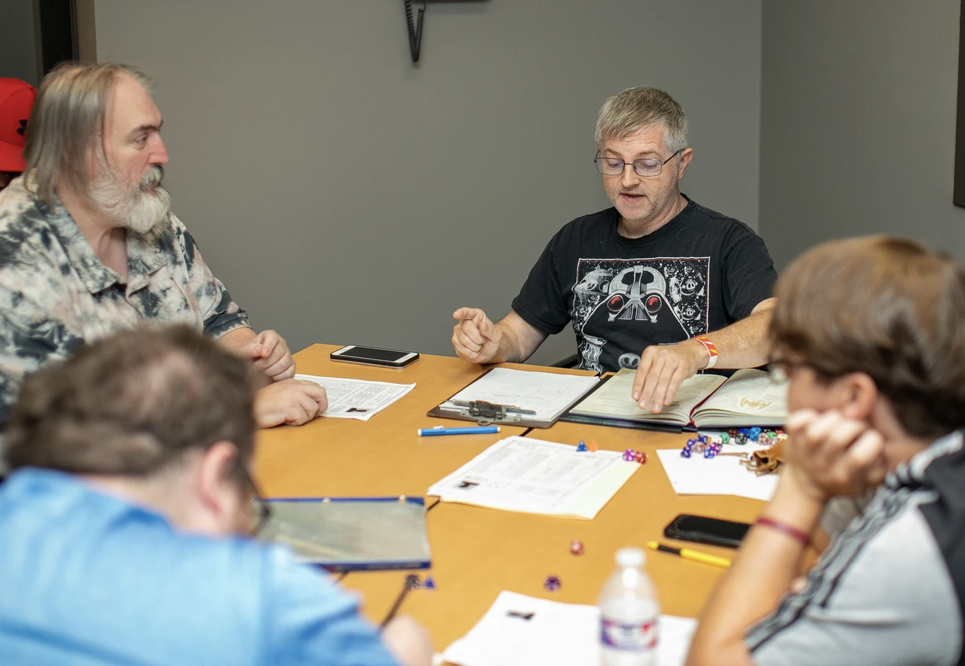 Five people sitting around a table engaged in a tabletop role-playing game, with character sheets, dice, and notebooks.