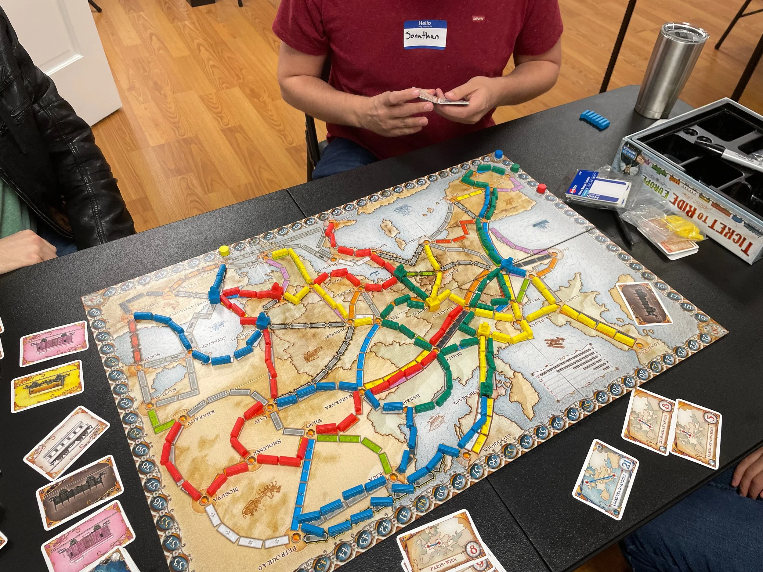 A group playing the board game Ticket to Ride, with colorful train pieces on a map of the United States. There are cards, a metal travel mug, and game pieces on the table.