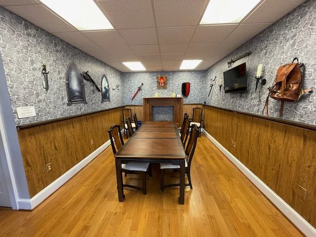 A rectangular wooden  table surrounded by eight chairs in a room with stone-textured walls and wood paneling, decorated with medieval-themed items including swords, a shield, and a backpack, with a fireplace, a flat-screen TV, and a small wall-mounted display on the far wall.