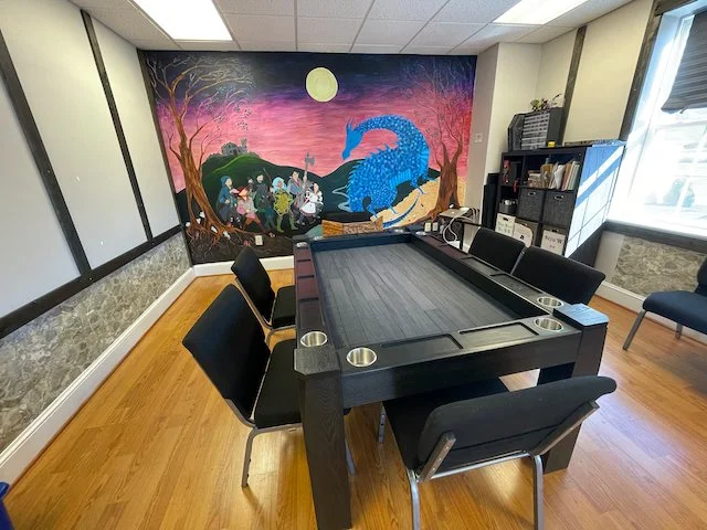 room with a large mural of a blue dragon and a group of people at night, outside a castle, with pink and purple sky and full moon. Six black chairs around a rectangular black table with built-in cup holders. Window on right side, bookshelf on the back wall.