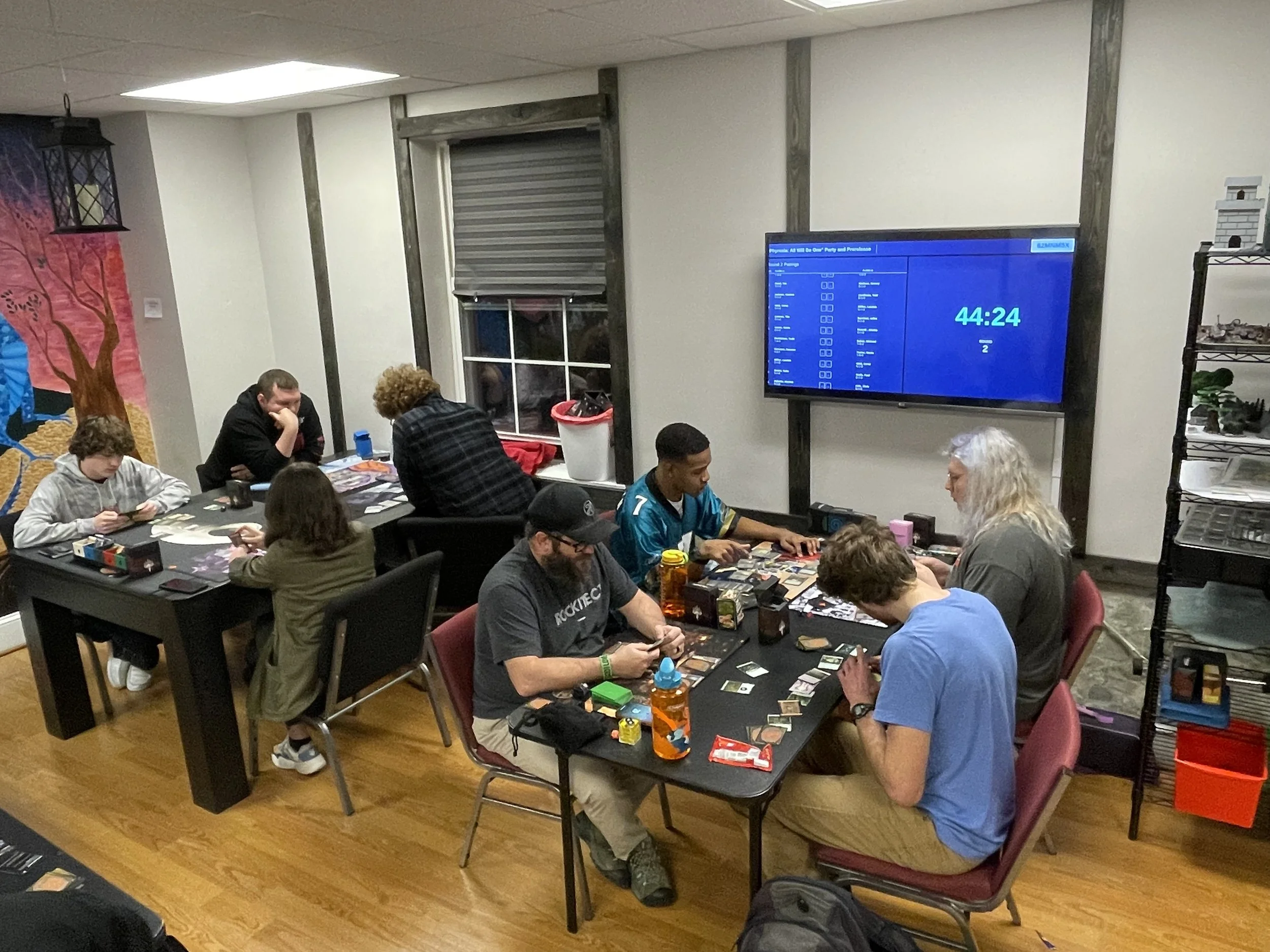People playing tabletop card games in a room with a large screen displaying a timer.