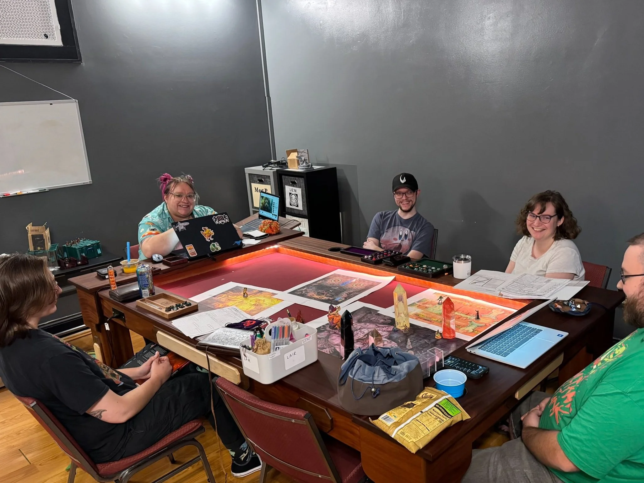 Group of five people sitting around a large table playing a tabletop role-playing game, with game maps, miniatures, drinks, snacks, and gaming accessories on the table. The room has a dark wall and a whiteboard on the wall.