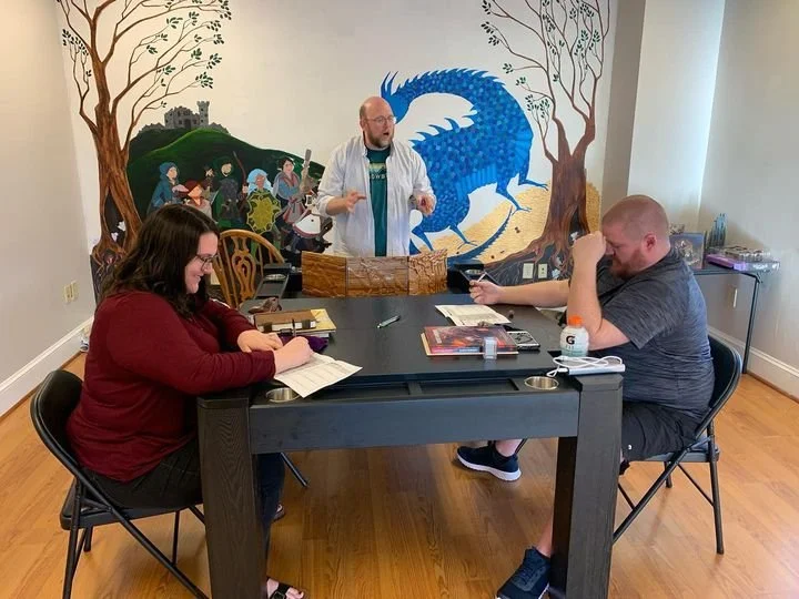 Three people sitting at a black table with books and a notepad, engaged in a discussion with a man standing, in a room with a large mural of a blue dragon and trees on the wall behind them.