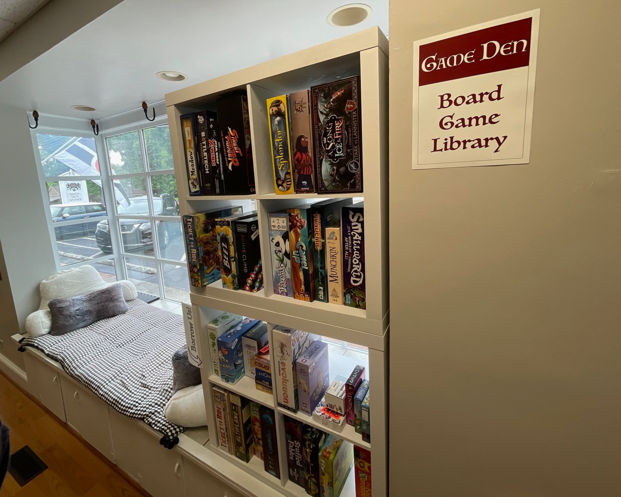 A bookshelf filled with board games in a game den, next to a window with a view of parked cars outside and a cozy window seat with pillows.
